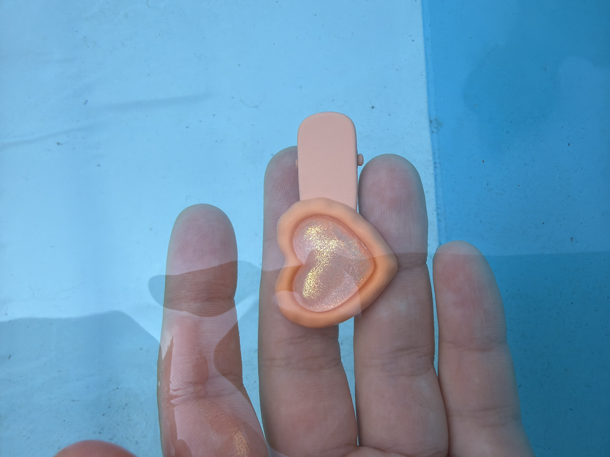 An overhead shot shows a hand holding a pink heart-shaped barrette in bright blue water. The water has some minor debris in it. The hand is submerged in the water, with skin texture clearly visible. The barrette has a glossy, glittery pink insert. The image appears to be taken in an outdoor swimming pool, possibly in the city of Qarağandı, Kazakhstan, given the context provided in the prompt, though there are no specific visual cues to confirm this location.