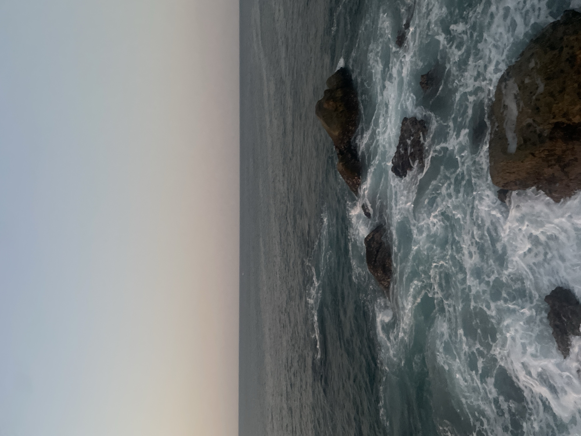 The image shows a portion of the coast in Puerto Escondido, Mexico. The scene features dark rocks battered by the ocean waves along the shoreline, with foaming white water surrounding them. The ocean extends to the horizon where it meets a pale sky, reflecting the ambient light.