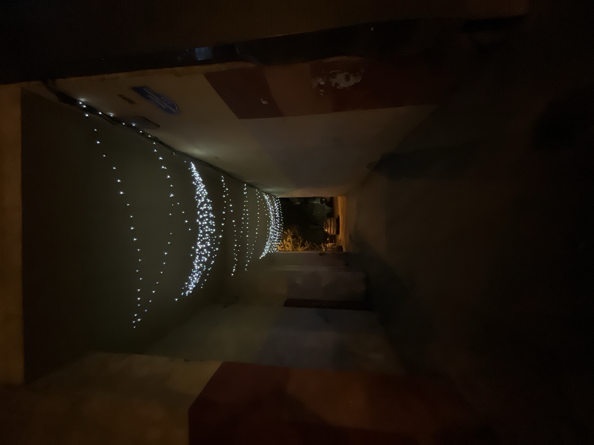 A narrow, dimly lit passageway or covered corridor is viewed from one end, looking towards a brighter opening at the far end. The overall scene is dark, indicating nighttime.
Along the upper left wall and part of the ceiling, numerous small, bright white string lights are strung, creating cascading, wavy patterns. These lights are the primary source of illumination within this section of the passage, casting a soft glow. On the ceiling, near the beginning of the passage, a small, rectangular blue sticker or label with faint, illegible white text is visible.
The right side of the passageway is notably darker. Closer to the viewer, a dark reddish-brown rectangular panel or surface is present on the lower part of the right wall. The floor of the passageway is mostly indistinct due to the low light.
At the distant end of the corridor, there is an opening that reveals a brighter, outdoor-like area. Through this opening, faint shapes of green foliage, possibly bushes or trees, and what appears to be part of a parked vehicle (perhaps a scooter or car) are discernable under ambient light. The string lights suggest a decorative or festive atmosphere within the otherwise utilitarian passage. No people are visible.