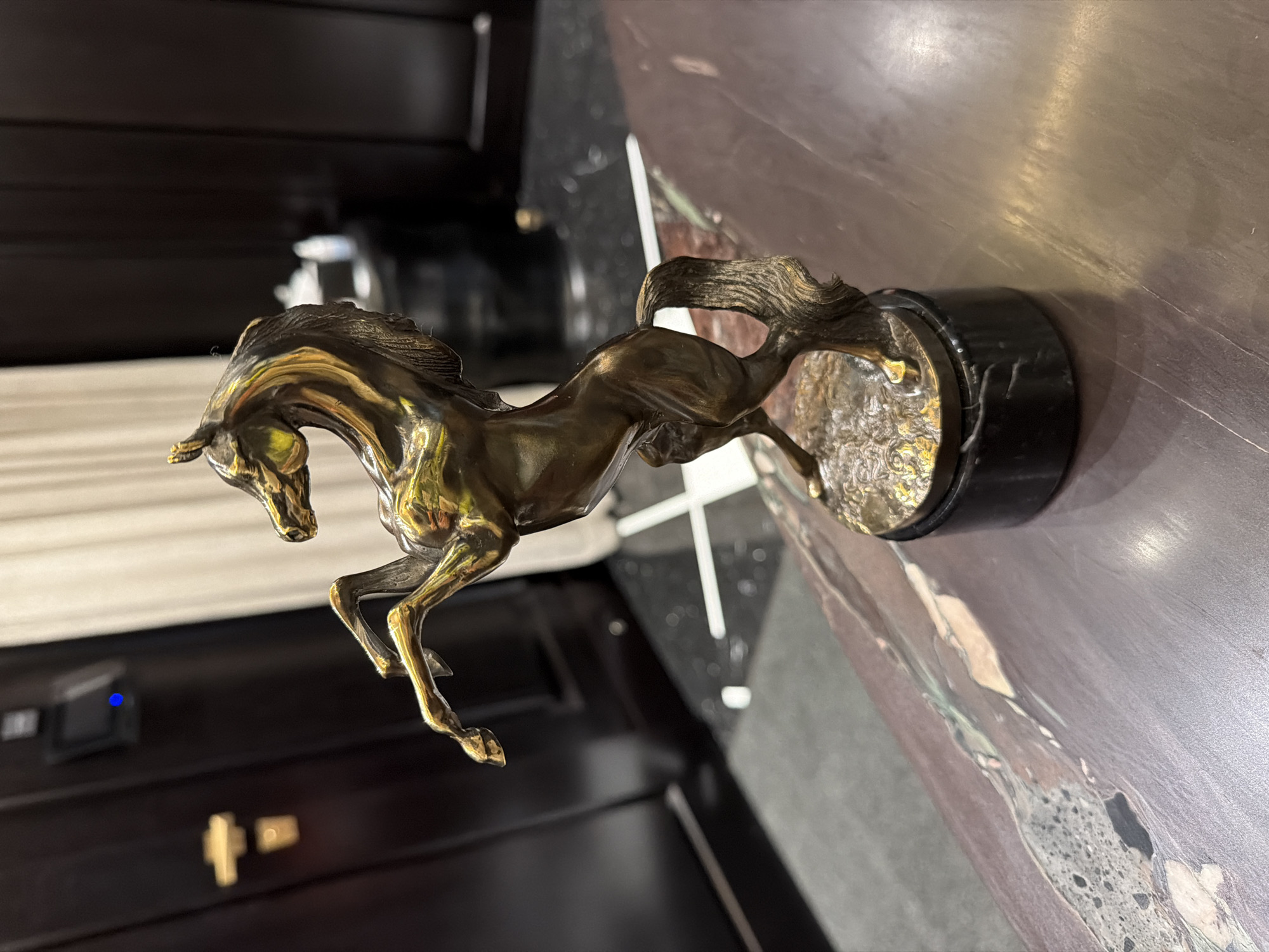 The image shows a close-up, angled shot of a metallic sculpture of a horse in motion, likely made of bronze or brass given its golden sheen. The horse is depicted mid-gallop, with its legs extended and mane and tail flowing. It is mounted on a dark, circular base, possibly marble. The sculpture is placed on a polished surface that appears to be a table or countertop, with a dark, reflective material underneath that shows some veining and texture.
In the background, there are hints of an indoor setting. A white curtain is visible to the left, and dark wooden paneling or furniture is present. There is also a small black electronic device with a blue indicator light on the dark surface. The lighting suggests an indoor environment, possibly a room with ambient light, as there are reflections on the polished surfaces. The scene appears still, with no people visible, and the focus is entirely on the horse sculpture. The overall impression is one of elegance and dynamism captured in metal.