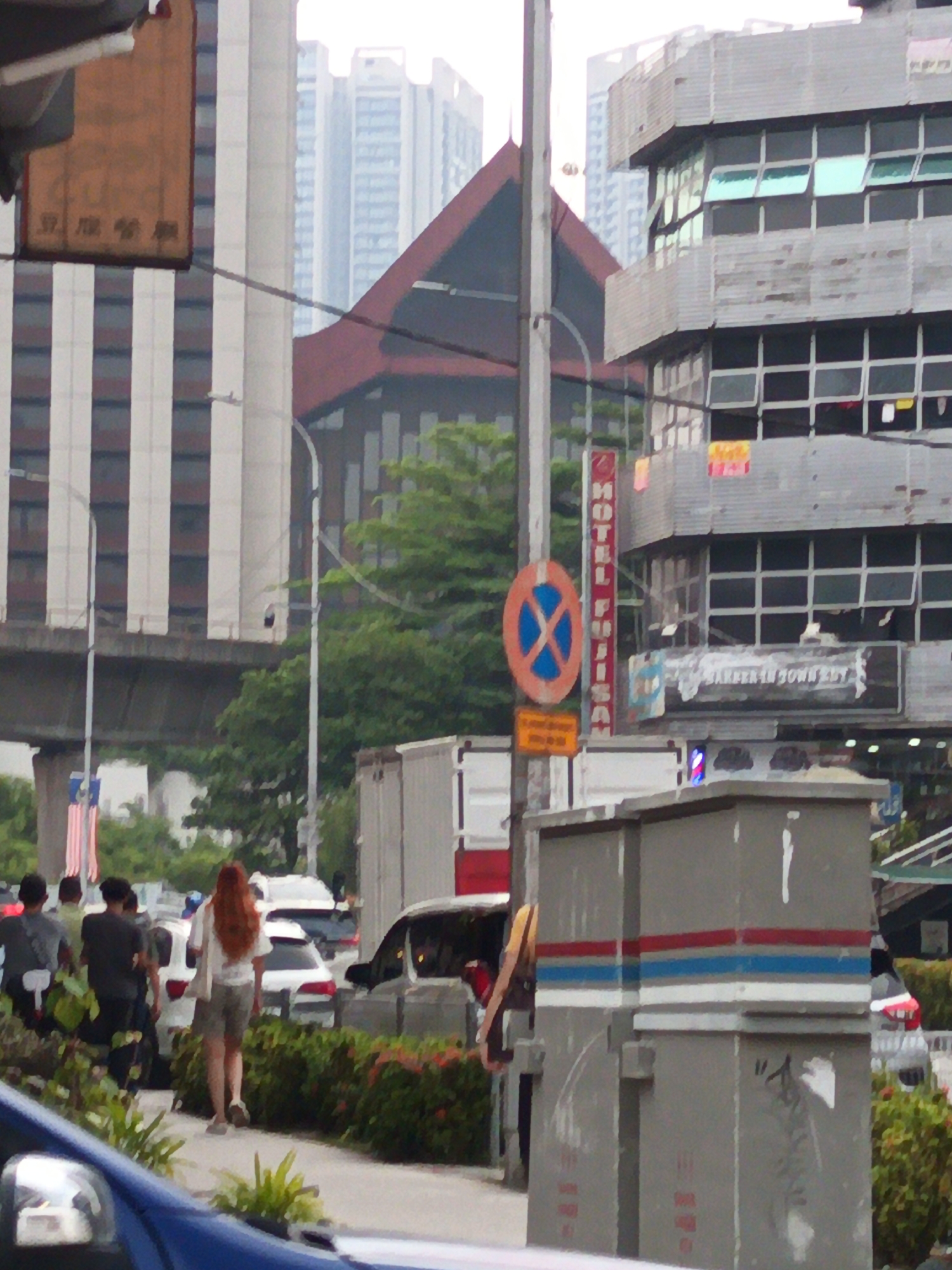 This is a medium-range, eye-level, street-level photograph taken in Kuala Lumpur, Malaysia, focusing on a busy urban intersection. The scene is dominated by multi-story buildings, including a modern glass-fronted structure and older, industrial-style buildings with exposed pipes and metal cladding. A prominent red and dark brown triangular-roofed structure with a cross-shaped window is visible behind a tall, grey utility box.
Several people are present. In the foreground, a woman with long, reddish-brown hair is walking away from the camera on a sidewalk. She is wearing a white t-shirt and grey shorts. To her left, a group of people are walking in the same direction; one person is wearing a dark shirt and has a backpack. Further back, a person in a grey shirt and dark pants is visible.
Traffic is present, with several cars and what appears to be a delivery truck. A blue car is partially visible in the bottom left corner of the frame. Traffic lights and street lamps are also visible. A red and white sign with a blue circle and a blue "X" inside it, indicating "no parking," is mounted on a pole. Another pole displays a vertical red sign with white text, possibly a hotel name. Malaysian flags are also visible on a pole in the background. The environment appears to be somewhat overcast.