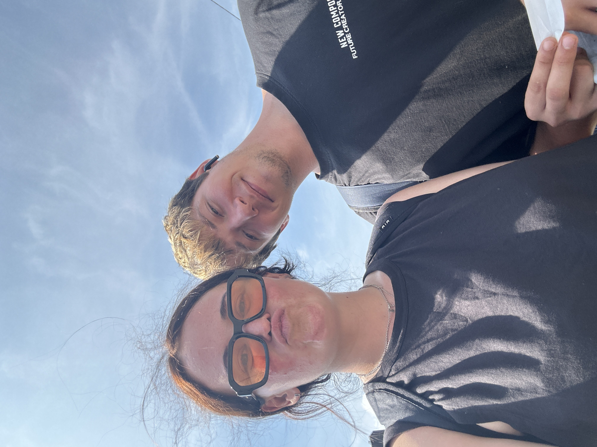 The image is a low-angle selfie of a young woman and a young man taken outdoors, with a bright blue sky and scattered clouds in the background. The woman is in the foreground on the left, pouting with her lips pursed and wearing large, orange-tinted square sunglasses. She has reddish-brown hair that is being blown by the wind. She is wearing a black t-shirt and a silver necklace. The young man is to her right, smiling and looking towards the camera. He has blond hair and is wearing a black t-shirt with white text that reads "NEW COMPOSURE FUTURE CREATOR". He has an earbud in his right ear. Both are wearing backpacks. The scene appears to be a sunny day, possibly in a public space. There is no specific indication of the city of Günzburg, Germany.