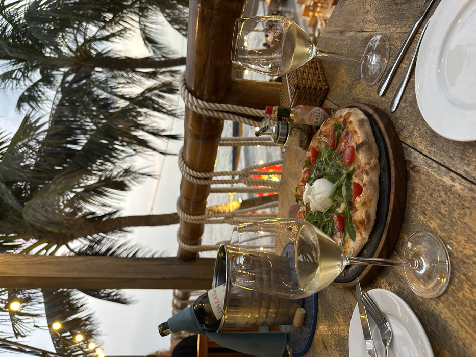A dining table is viewed from a slightly elevated perspective in an outdoor restaurant setting in Thành phố Phan Thiết, Vietnam, at dusk. In the immediate foreground, on a rustic wooden plank table, a pizza with burrata, arugula, and cherry tomatoes rests on a dark round plate atop a wooden serving board. To the left of the pizza, two white wine glasses are positioned, one full and closer to the viewer, the other partially visible on the right. A chrome ice bucket containing a green-wrapped "DALAT" branded wine bottle sits on a blue and white coaster to the left of the wine glasses. Cutlery and a white plate are placed to the far left. To the right of the pizza, a small bottle of Tabasco sauce and a clear glass condiment dispenser with ground spices are present next to a small woven basket.
The midground features a prominent wooden railing adorned with thick white rope, creating a nautical theme. Beyond this railing, the dark expanse of the sea or ocean is visible. The background reveals multiple tall palm trees silhouetted against a pale sky, indicating twilight. String lights with visible bulbs are strung above, illuminating the dining area. Faint, warm lights are visible in the distant lower background, suggesting another section of the restaurant or a nearby area at a lower elevation. A single individual is partially visible sitting at a table in the far left background.