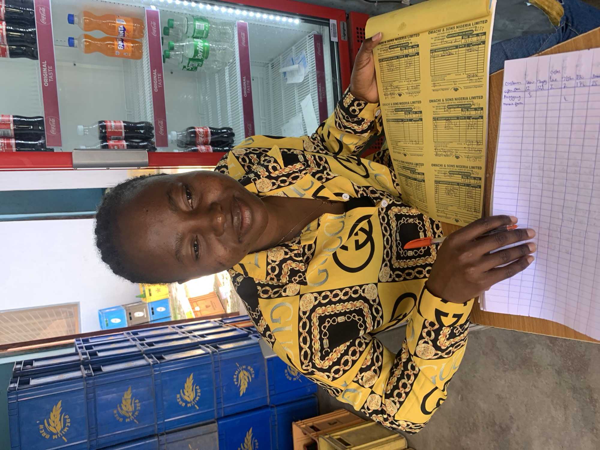 The image depicts a woman in a brightly colored yellow shirt with a black and gold chain pattern, sitting at a wooden desk. She is looking down at a yellow ledger or notebook open on the desk, which is covered with handwritten entries and columns. Her right hand is resting on the ledger, and she is holding a red pen with her index finger. In front of the ledger is a white notebook with lined paper, also containing handwritten notes.
Behind the woman, to her left, are stacked blue plastic crates with yellow lettering that reads "BREW PREMIUM." To her right is a red, upright refrigerator displaying bottles of Coca-Cola and Fanta on shelves. The refrigerator has a glass door with visible interior lighting.
The environment appears to be an indoor space, possibly a shop or store, given the presence of goods like beverages and crates. The backdrop includes a portion of a wall with some windows or ventilation. The overall impression is one of a business setting where transactions or inventory are being managed. The scene is well-lit, suggesting an operational environment.