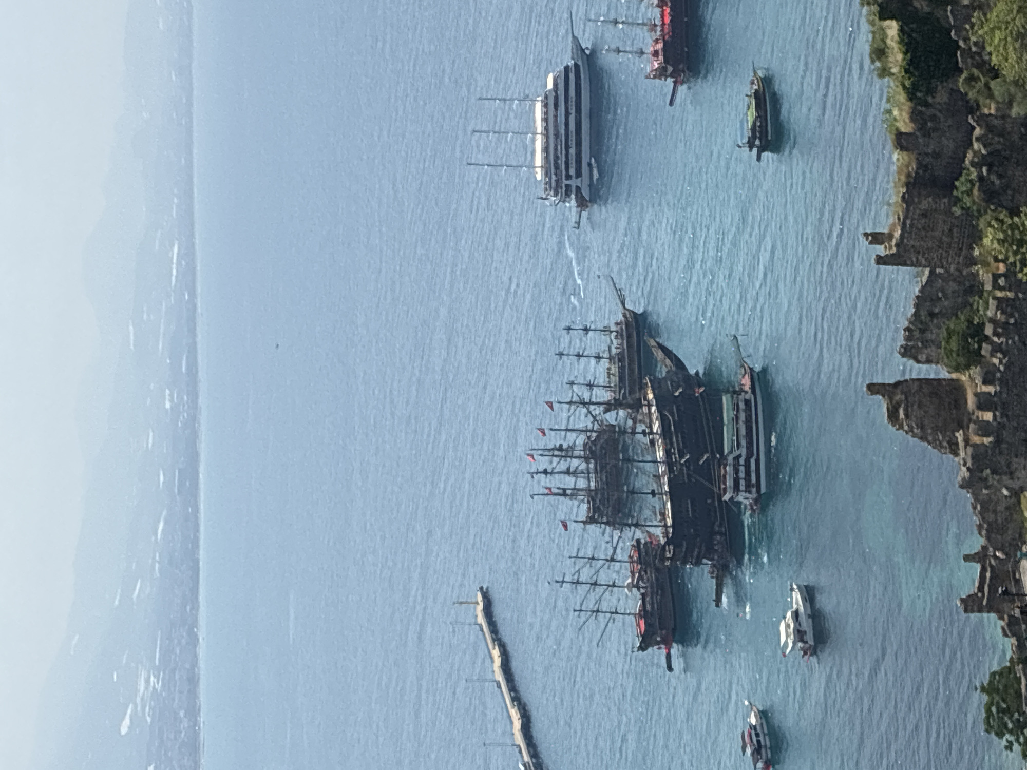 The image displays a panoramic view of the Alanya harbor in Turkey, with the Alanya Castle ruins in the foreground. The harbor is filled with various vessels, including several large, traditional wooden pirate ships with multiple masts and red flags, a modern, multi-decked white passenger ship, and smaller motorboats. The water is a clear turquoise, and the background shows the hazy outline of mountains and the coastline of Alanya. The overall scene captures a bustling maritime activity within a historic coastal setting.
