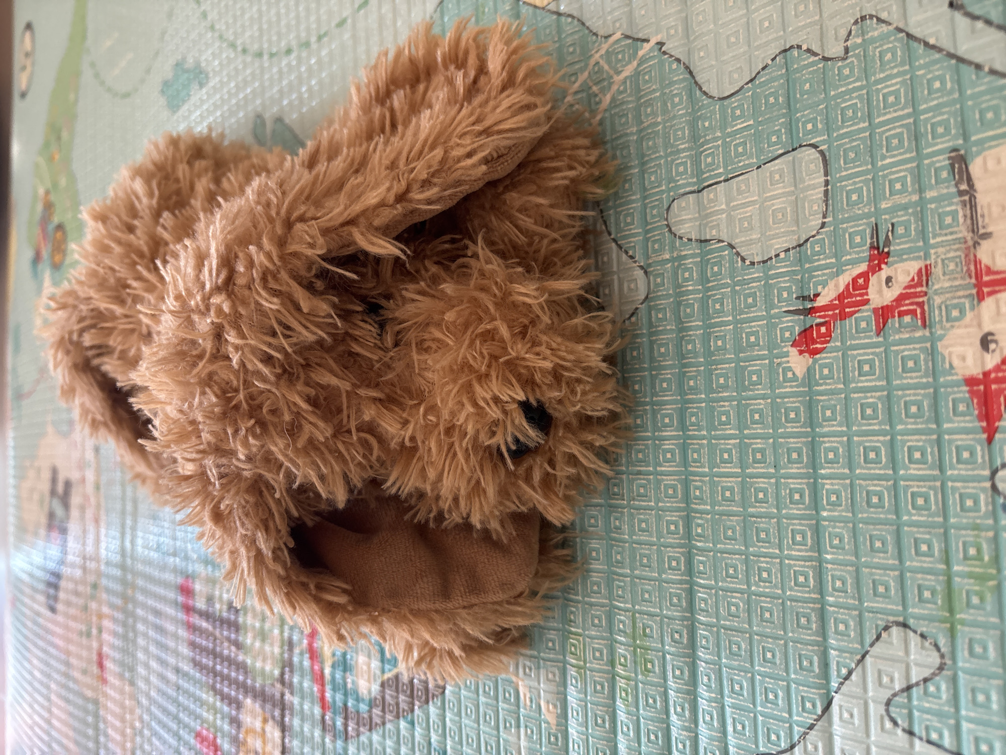 A brown, shaggy-haired plush toy, resembling a canine or ursine effigy, is depicted in a prone position on a textured playmat. The toy's synthetic fur is a medium brown color, and two small, black, spherical eyes are visible on its partially obscured face. Its floppy ears, made of a smoother, lighter brown fabric, frame its head. The plush toy is situated on a light blue-green polymeric playmat featuring a repeating embossed pattern of small squares. Portions of graphic illustrations are faintly visible on the mat, including a red and white airplane motif in the lower left section and irregular black outlines that suggest a map or diagram. The background consists of a blurred extension of the same patterned playmat. The scene is recorded within a residential or recreational environment in the city of Dubai, United Arab Emirates.
