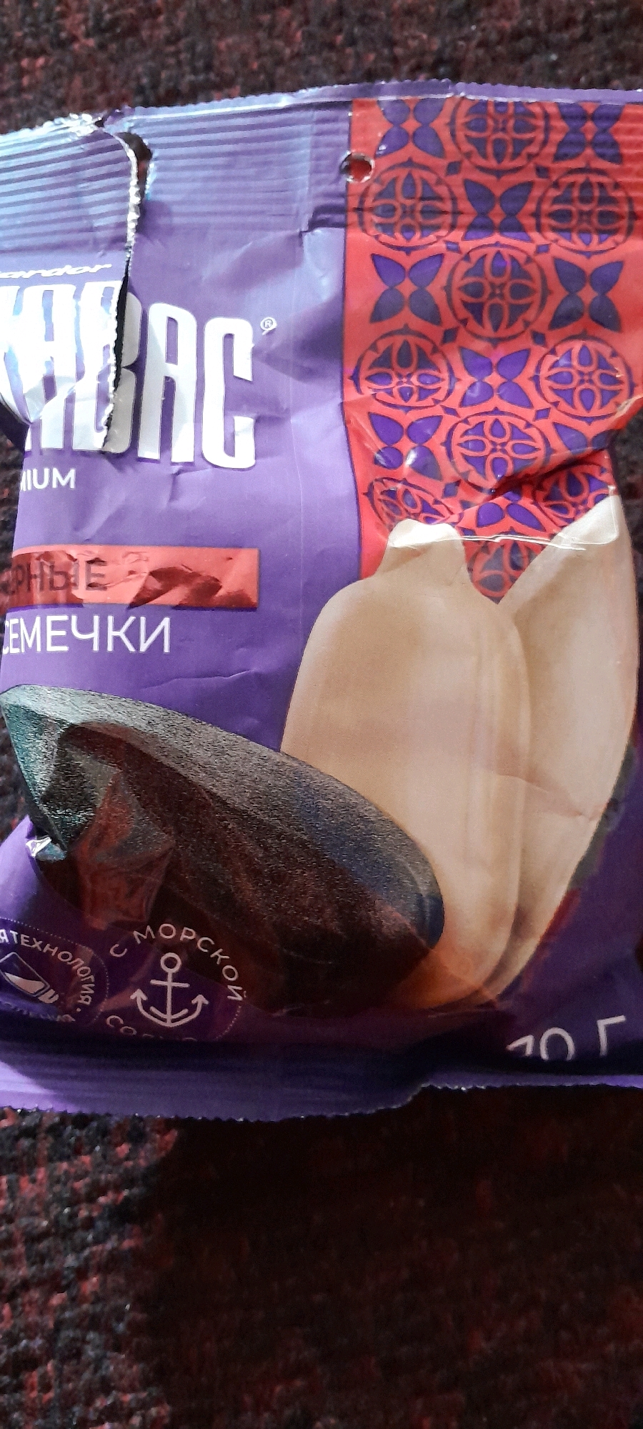 A torn bag of sunflower seeds sits on a red and black textured surface, possibly a carpet or upholstery. The bag is purple and features a red and blue patterned design. A large, dark sunflower seed is prominently displayed on the front of the bag. Text on the bag is partially visible, including "UM" and "СЕМЕЧКИ" (which translates to "seeds" in Russian). A circular emblem with an anchor is also visible. The image is a close-up, with no indication of its location in Отқаноқ, Tajikistan, or any surrounding environment.