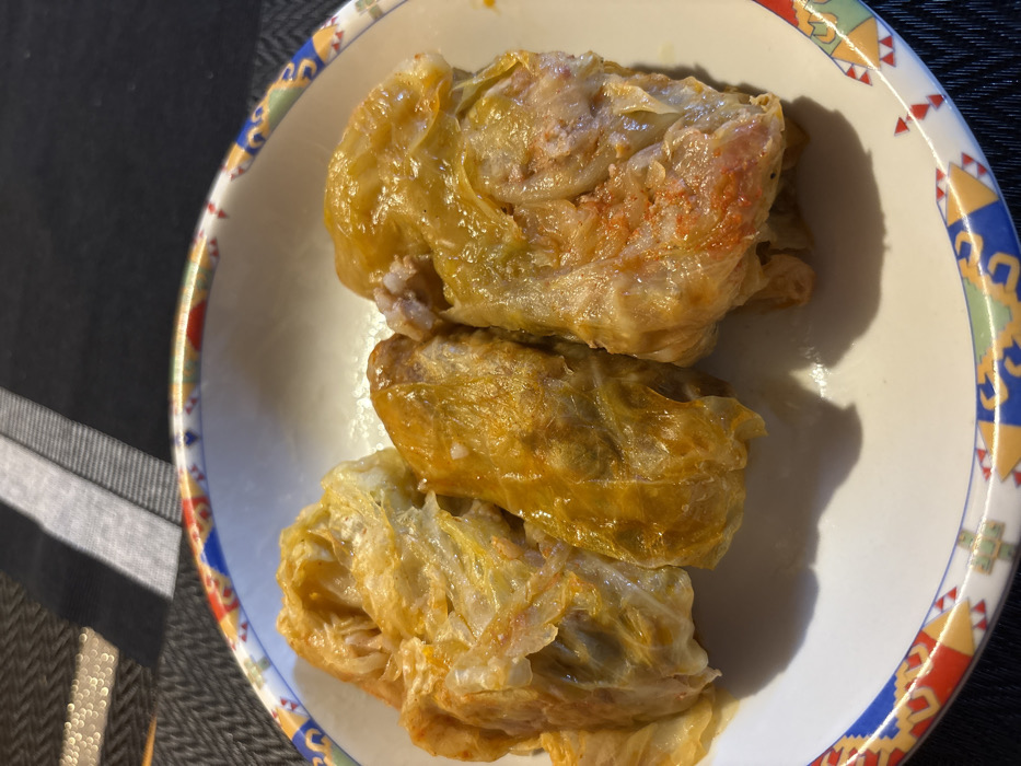 The image presents three portions of cooked cabbage rolls, likely sarma or golubtsi, arranged in a white, shallow ceramic bowl. Each roll displays a yellowish-orange color, indicative of its cooked state, with some areas of the cabbage leaves appearing lighter. The rightmost roll reveals a reddish-pink meat and rice filling through a partial opening in its leaf. The bowl features a distinctive rim adorned with a multi-colored pattern incorporating blue, red, yellow, and green geometric and folk-art inspired motifs. The bowl is situated on a dark-colored placemat or table runner with a visible woven texture, possibly herringbone, and is set against a background featuring light gray and black stripes. This scene is located in the city of Stefan Karadja, Bulgaria.