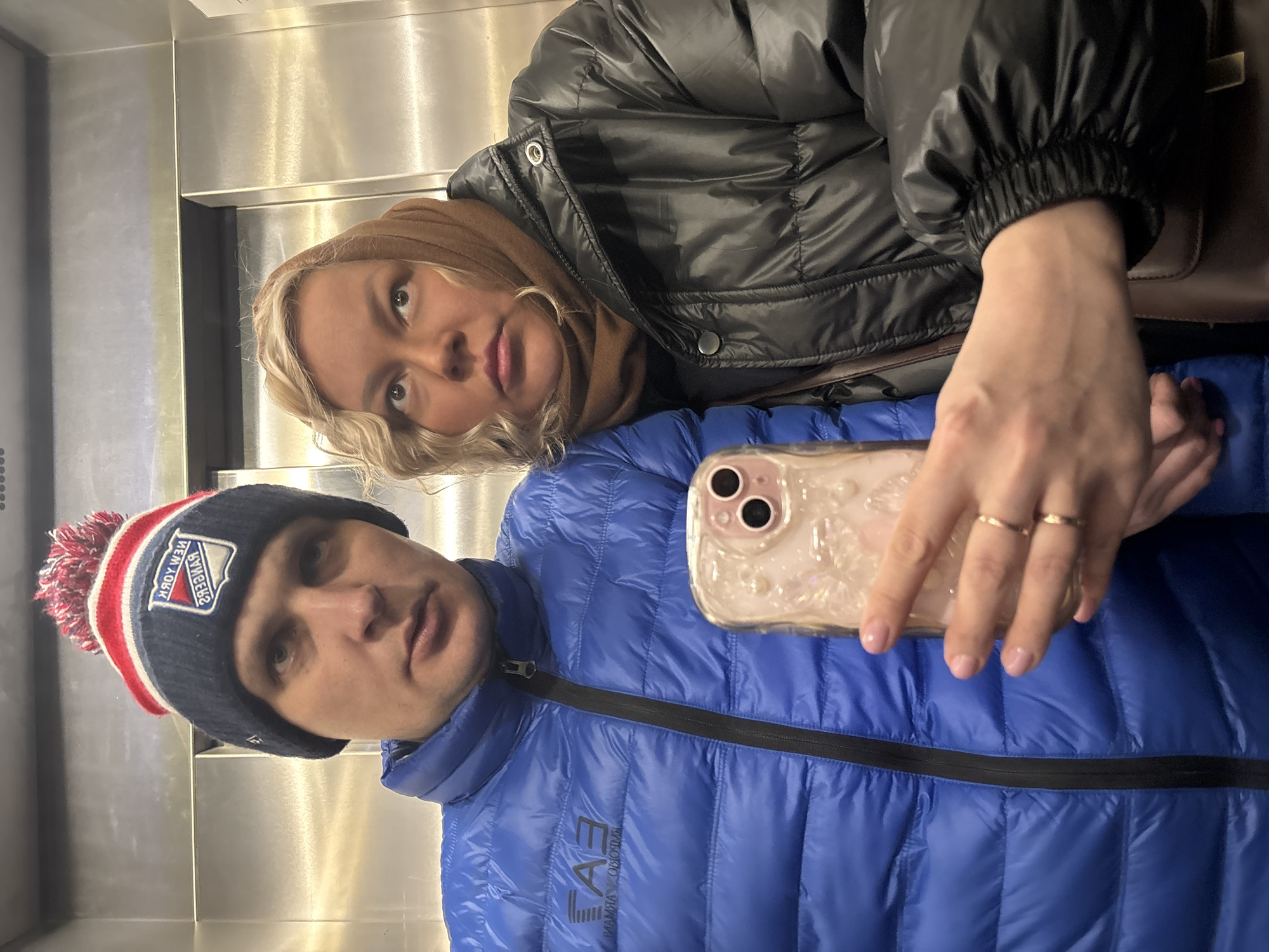 A blonde woman with light skin wearing a black puffer jacket and brown scarf takes a selfie with a man wearing a blue puffer jacket and a New York Rangers beanie. The photo is taken inside an elevator with metallic walls. I cannot verify the location in Izhevsk, Russia, based on the information provided.