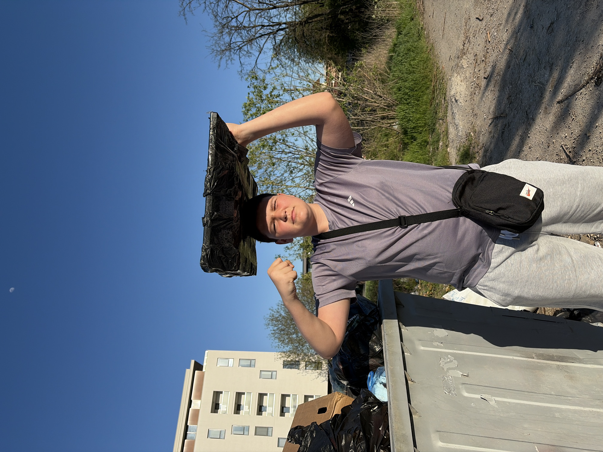 A young male stands at the edge of an overflowing trash container, positioned outdoors in what appears to be an urban or suburban area. The male wears a t-shirt, sweatpants, and a crossbody bag. He holds a large, black object, possibly a container lid, over his head. The container is filled with black trash bags, cardboard, and other waste. Behind the male is a building with multiple windows, and beyond that is an area with trees and vegetation. The sky is a clear blue, with a visible moon in the upper left corner. The exact location in the city of Sofia, Bulgaria cannot be determined from the image alone.