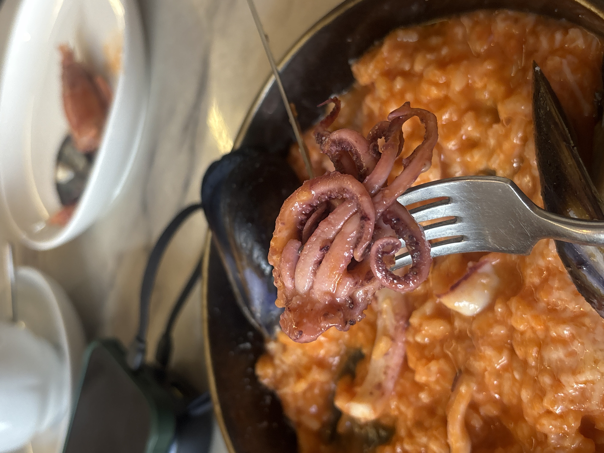 A close-up shot depicts a fork holding a small cooked octopus, likely part of a seafood dish. The octopus is reddish-brown and shows curled tentacles. Behind the fork, there appears to be a dish of orange-colored risotto with mussels. There's also a smaller, white bowl visible in the background, seemingly containing another seafood item. The setting is likely a restaurant. The image does not contain any evidence to confirm the location in Moscow, Russia.