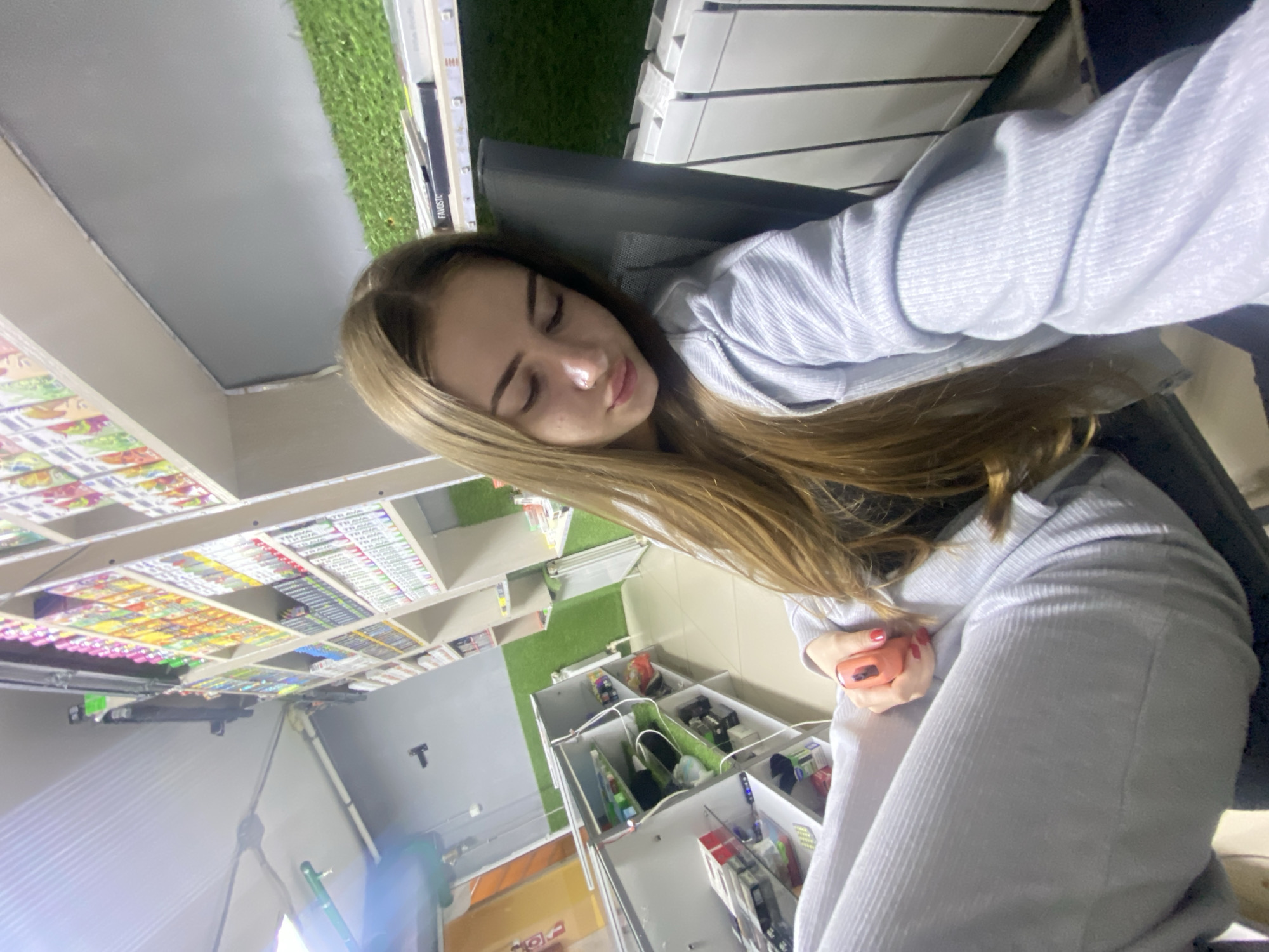A young woman is depicted taking a selfie inside a retail store in the city of Дьяково, Russia. She has long, light brown hair and is wearing a light-colored, ribbed long-sleeved top and matching pants. Seated in a dark chair, she is looking downwards with her eyes partially closed. Her left hand, adorned with red nail polish, holds a small, orange, curved object. The background reveals rows of shelving units stocked with numerous brightly colored packaged goods, suggesting a specialty store. Walls are partially covered with green artificial turf panels, and white radiators are visible on the right. Overhead lighting illuminates the scene. A label reading "FAVOSTIC" is visible on a shelf.