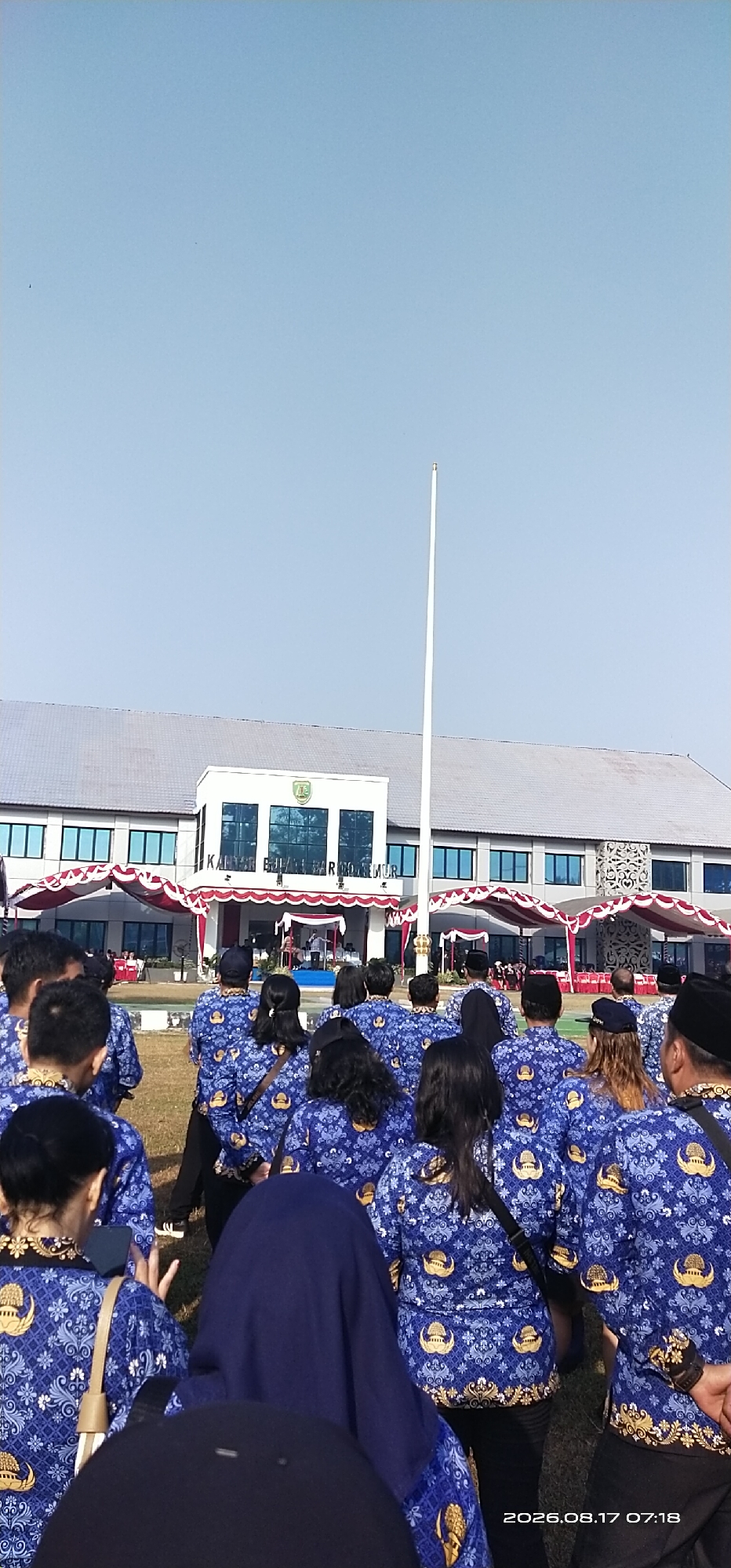 The image depicts a group of people, mostly women, wearing matching blue shirts with a gold emblem, standing in what appears to be an outdoor assembly. They are facing away from the camera, towards a building. The building is a large, modern structure with numerous windows and a gray roof. In front of the building, there is a stage or platform where several individuals are visible, seemingly engaged in a ceremony or presentation. Red and white decorative awnings are erected in front of the building. A tall, white flagpole stands prominently to the right of the building. The ground in front of the building is covered in grass, and there is a clear blue sky above. The scene is likely taking place in the city of Tamiang Layang, Indonesia, as suggested by the text overlay "2026.08.17 07:18" which, though a future date, is a common format for date and time stamps in Indonesian contexts. The people are predominantly wearing uniforms, suggesting a formal gathering, possibly a government or official event.