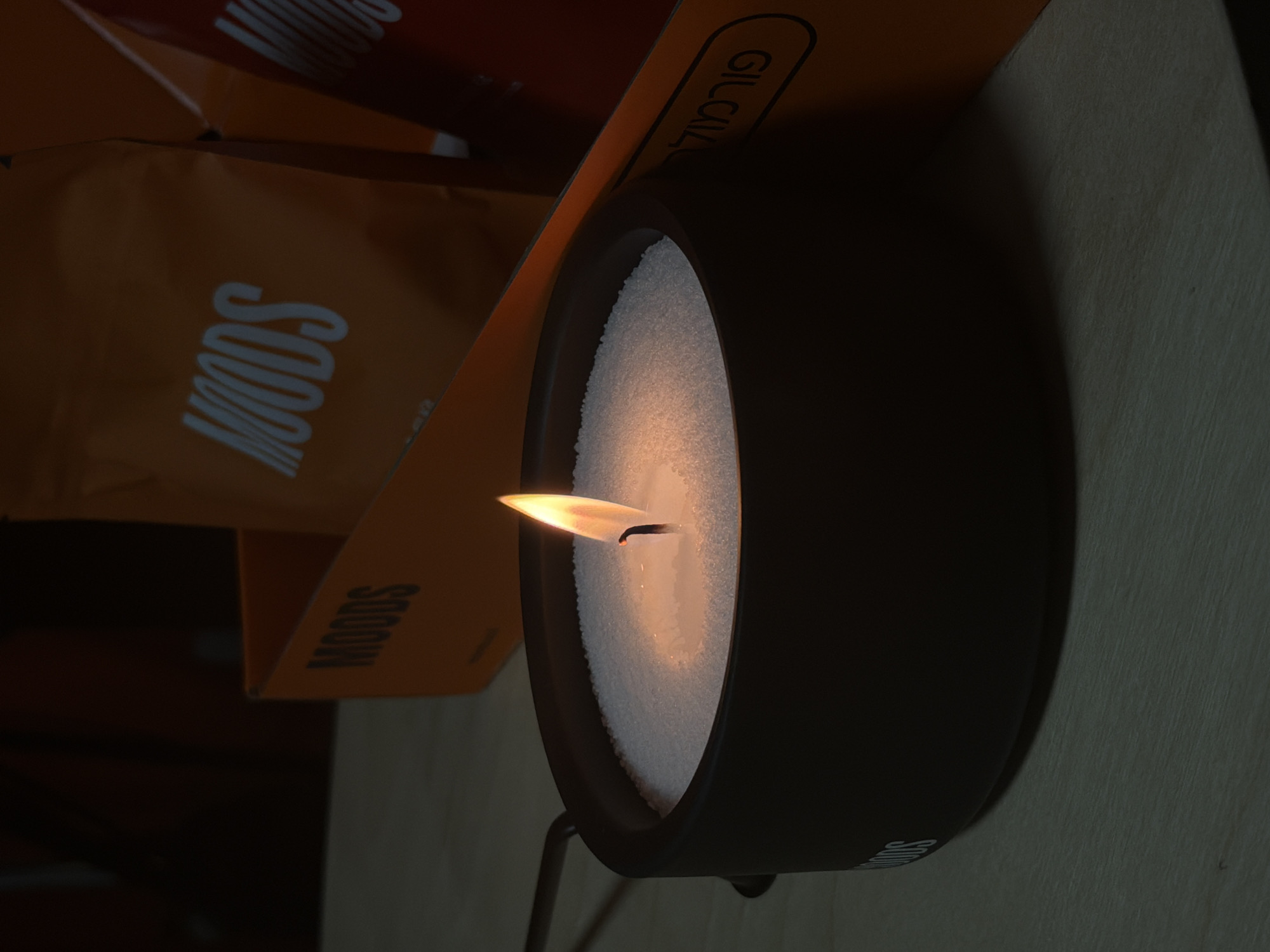 An indoor scene, possibly in Ashkelon, Israel, features a lit candle in a dark, round container filled with white granular material. The candle flame is bright orange and flickers. In the background, an orange cardboard box with the word "MOODS" printed in white is visible. To the right of the orange box is a portion of a red object, also with "MOODS" printed in white. A portion of text in Hebrew is visible on the orange box, reading "פורמול" (formul). The objects are placed on a light-colored wooden surface.