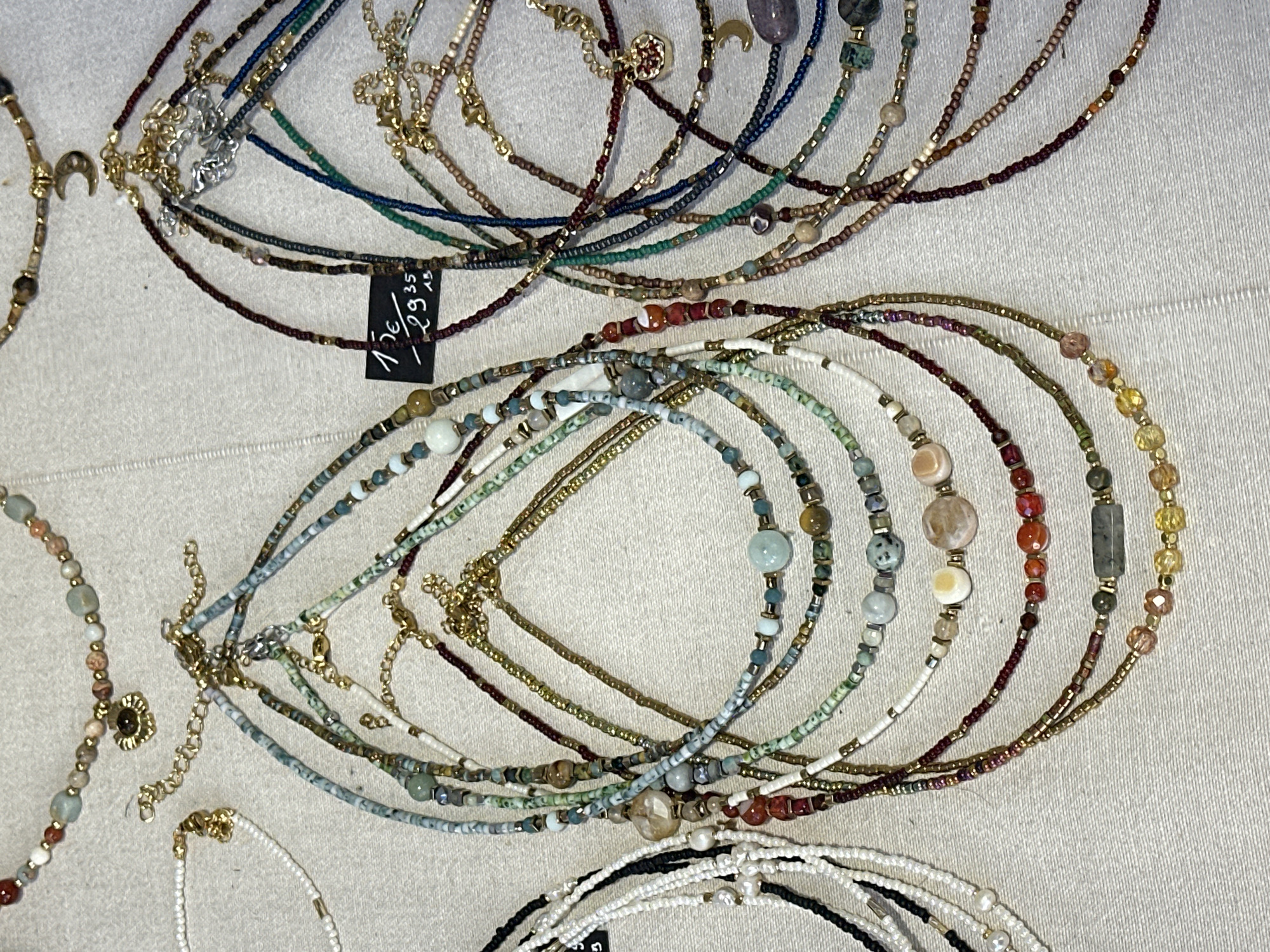 The image shows a display of handmade beaded necklaces on a light-colored surface. The necklaces are various colors and styles, including gold, silver, red, white, blue, and green. One of the necklaces has a price tag that reads "136 / 29,35". The location is Sofia, Bulgaria.