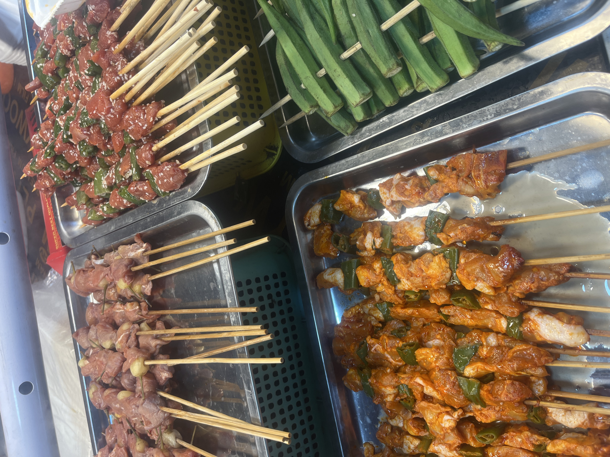 The image depicts an outdoor food stall in Da Nang, Vietnam, showcasing an array of skewers ready for grilling. In the foreground, a rectangular metal tray holds numerous skewers of marinated chicken and green peppers, coated in a reddish-orange spice mix. To the right, another metal tray is filled with whole okra pods, also skewered. Behind these, two more trays are visible. The tray on the left contains skewers of raw meat, possibly beef or lamb, interspersed with small, light-colored round pieces, possibly garlic or onion. The tray on the right holds similar raw meat skewers, these ones adorned with green pepper pieces and sprinkled with sesame seeds. The food stall appears to be part of a larger market or street food area, with various containers and food items visible in the background. The lighting suggests it is daytime.