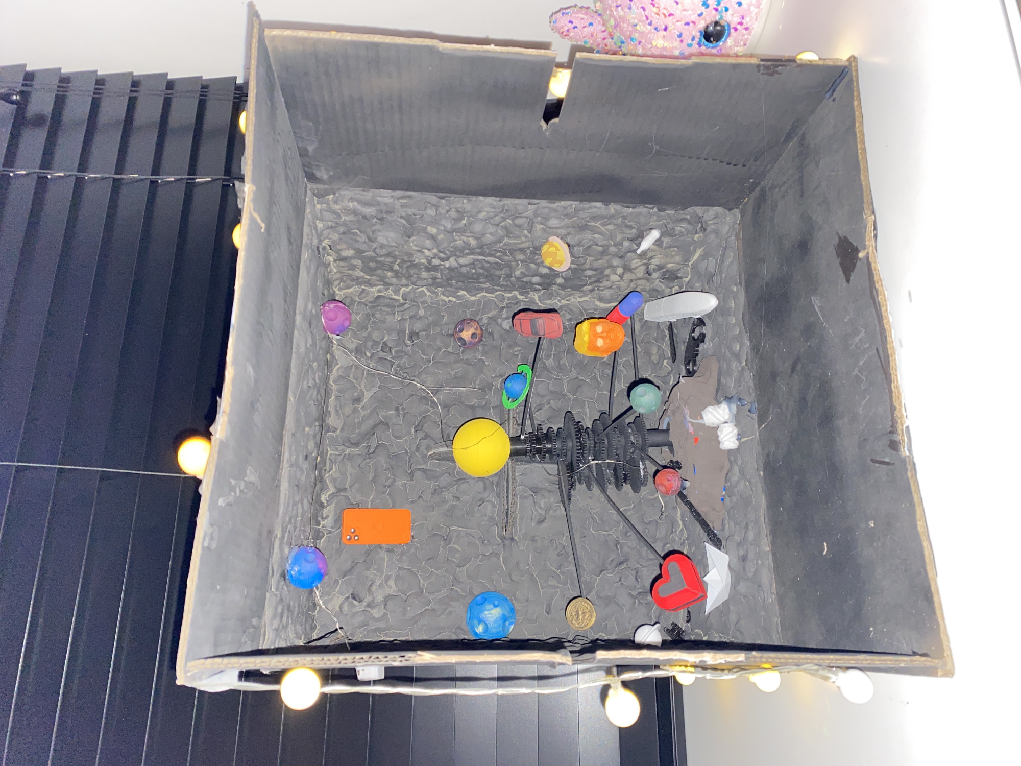 A top-down view reveals a dark, gray cardboard box containing a model of a solar system or similar cosmic scene. Various colorful, spherical objects are arranged on a gray, textured surface within the box. A central, gear-like structure anchors these objects, suggesting a mechanism or movement system. The box's interior surface is finished in a cracked or marbled pattern. String lights are positioned around the box's exterior. The image provides no information about location.