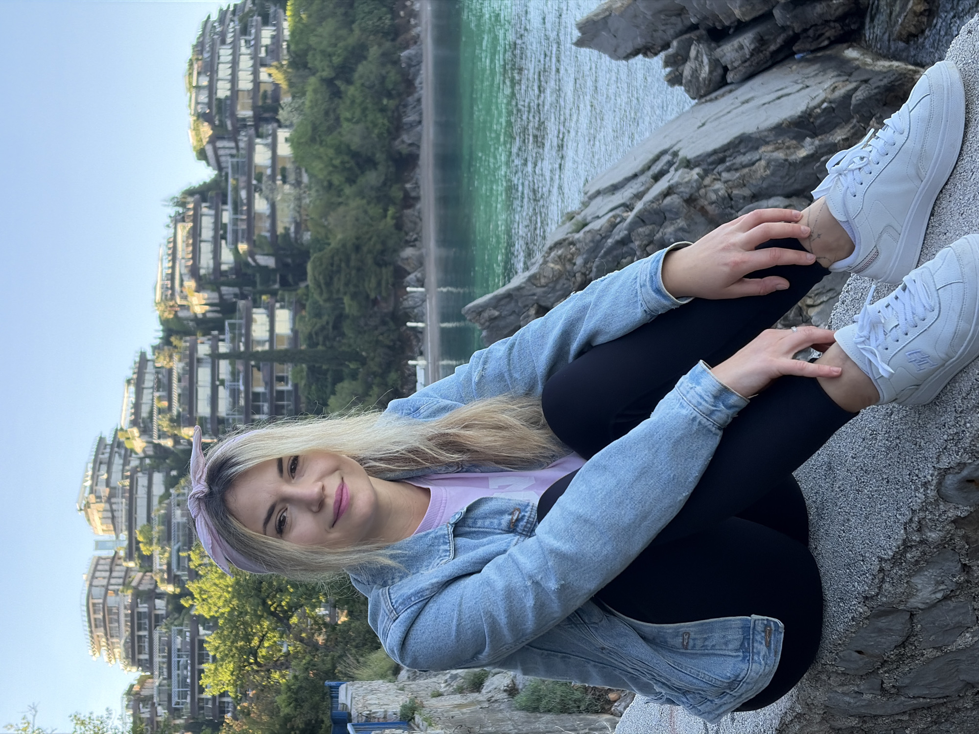 A young woman is sitting on a rocky outcrop with a view overlooking the Adriatic Sea. Her appearance suggests she is in her late teens to early twenties. She is wearing black leggings, a light-colored top, a denim jacket, and white sneakers. She also has a light purple headband. The background features tiered buildings nestled into a hillside, characteristic of the architecture in Budva, Montenegro. The water in the foreground is calm and appears to be a light turquoise color. The sky is clear and bright. The image is shot outdoors in daylight, likely in the early morning or late afternoon given the soft light.