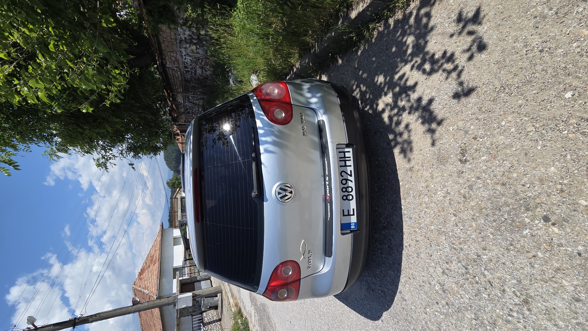 A silver Volkswagen Golf hatchback, identified by Bulgarian license plate 'BG E 8892 HH' (Lovech Province), is parked facing away from the viewer on a gravelly asphalt road. The vehicle's rear displays 'Golf' badging with an Ichthus symbol on the left, '2.0 TDI' badging on the right, and a 'sports' decal applied to the black bumper protector. The rear window is tinted, reflecting bright sunlight, and tree shadows are cast across the vehicle's right side and the road. To the left, a concrete utility pole supports multiple overhead lines, positioned in front of a white house with a terracotta tiled roof. To the right, a large leafy green tree overhangs a stone and brick wall, with tall green grass growing along the road's edge. Distant hills are visible on the horizon beneath a blue sky with scattered white clouds.