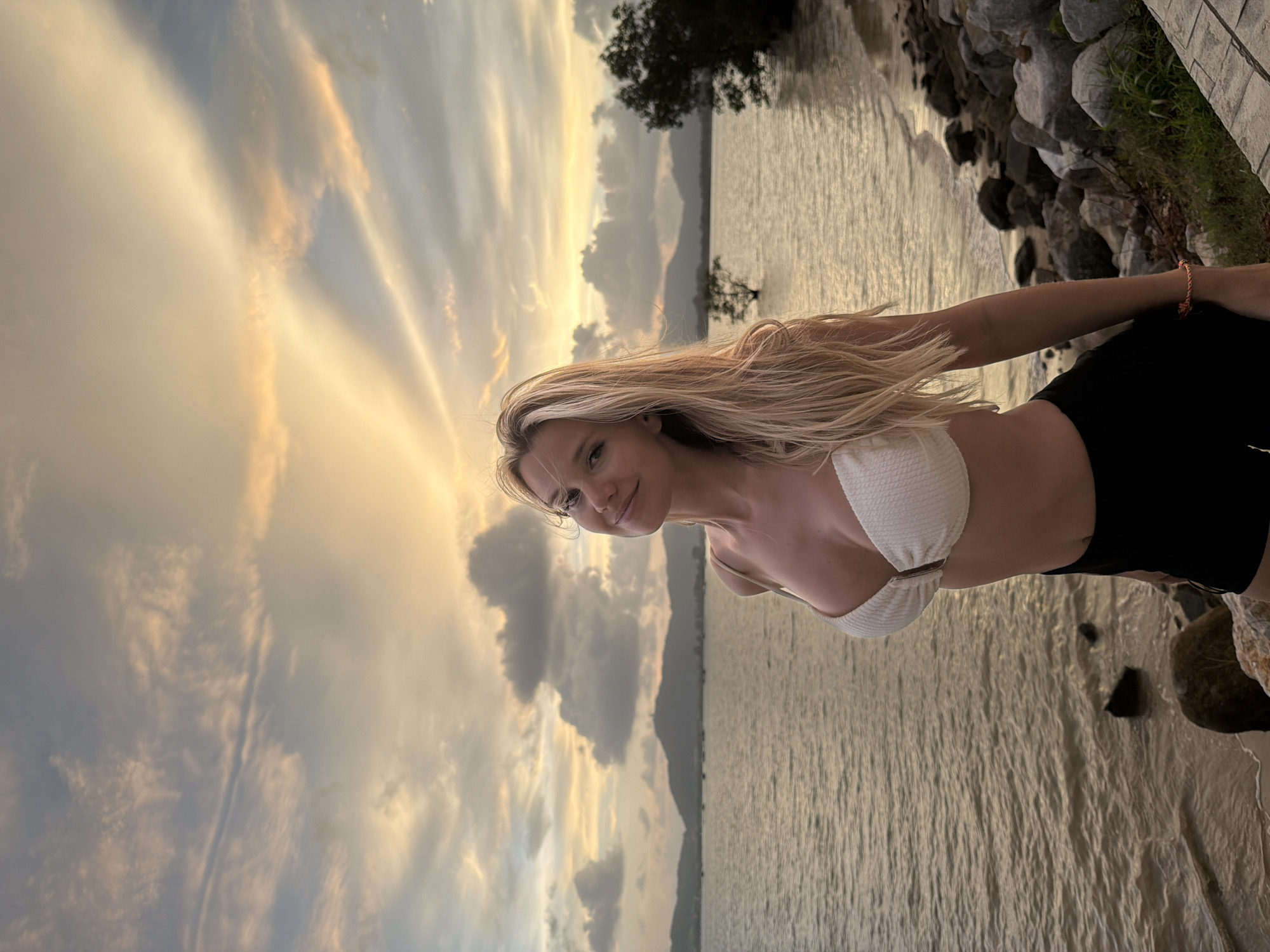 A young blonde woman wearing a white bikini top and black shorts stands on a rocky shore in Chalong, Thailand. The water of the ocean is calm and reflects the colors of the cloudy sky, which is illuminated by a sunset. In the distance, mountains can be seen, and a solitary tree stands in the water. The woman is looking at the camera and smiling. The overall impression is one of serenity and natural beauty.