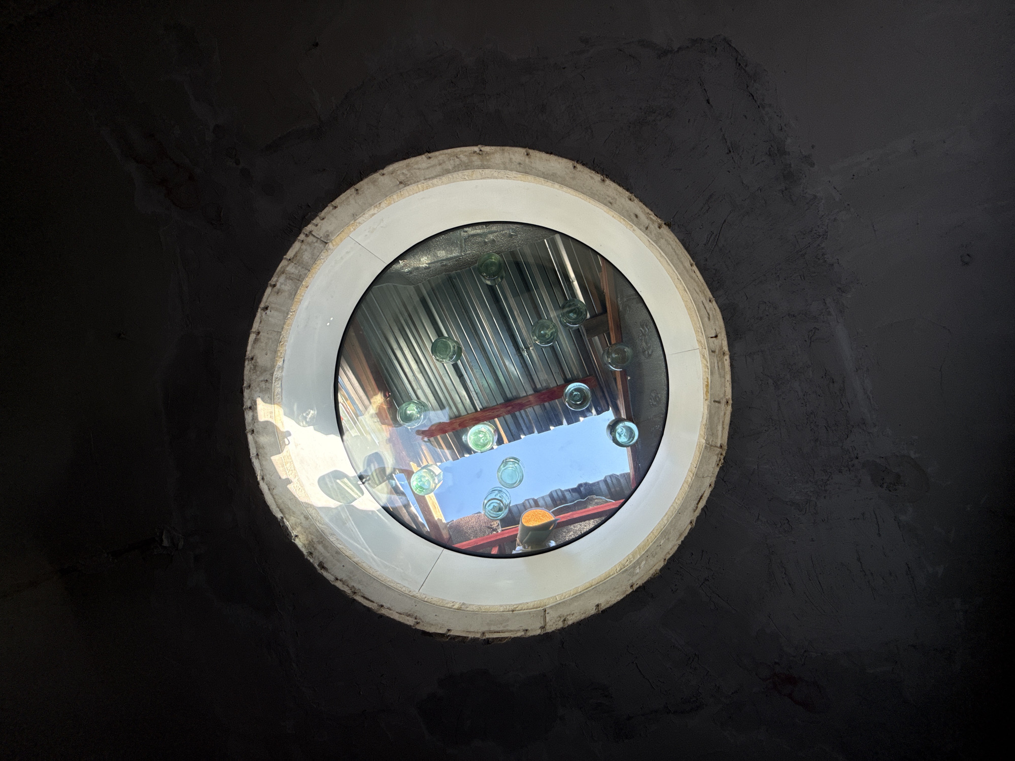 A round window, possibly a skylight, is set into a dark, textured ceiling, likely made of concrete or plaster. The window frame appears to be constructed of unfinished wood and white material. Through the glass, a reflection or view shows a corrugated metal roof structure, beams, and several small, round, greenish glass objects. Above this, a clear blue sky is visible, with some reddish-orange elements that could be part of a building or an object. The lighting suggests daylight. The image does not contain any identifiable people or actions. Based on the visual elements, such as the corrugated metal and the style of construction, this scene is plausible for a creative or industrial space, which can be found in various parts of Moscow, Russia, including renovated factory districts or art studios.