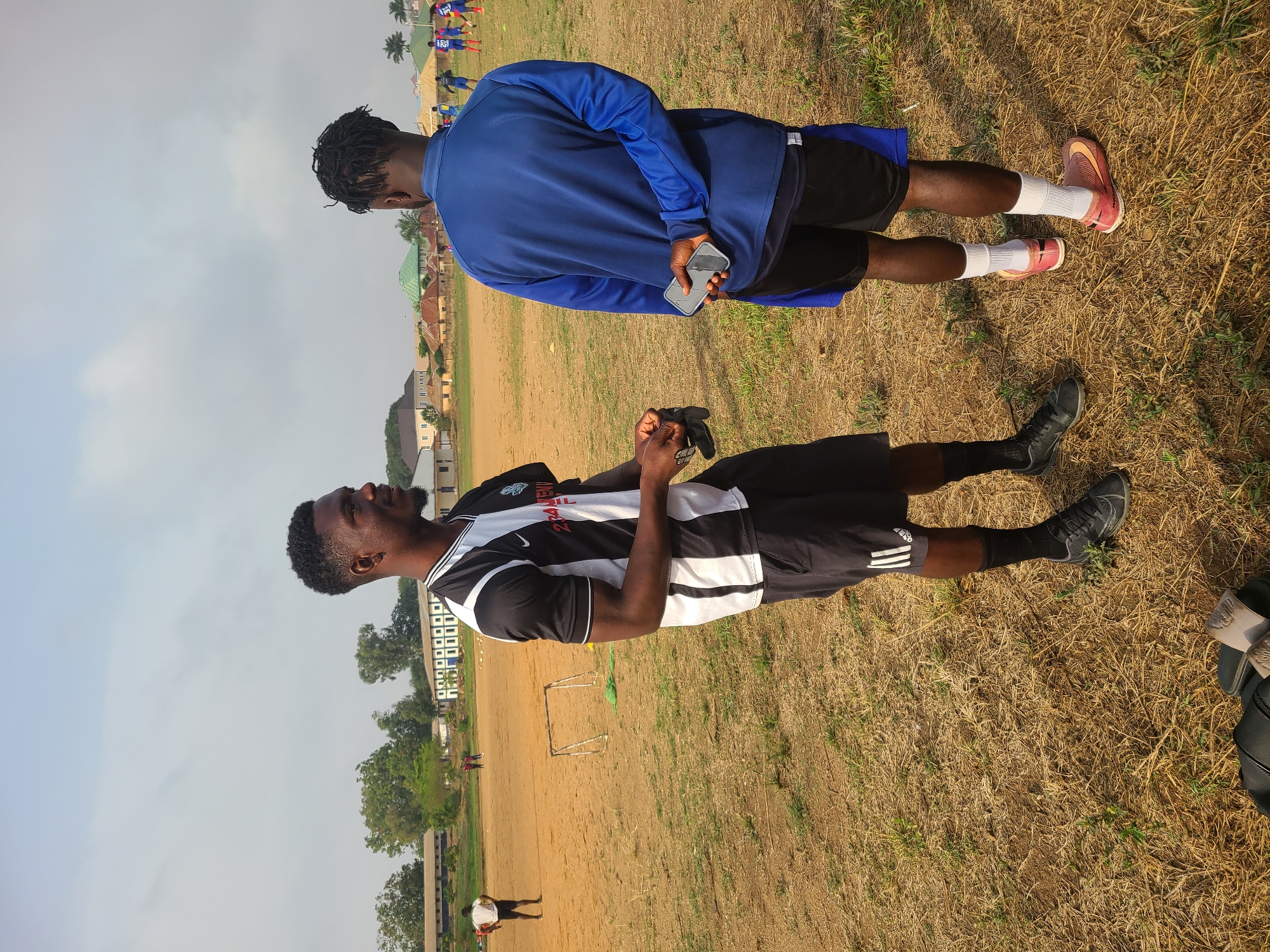 Two young men stand on dry grassy ground in Bukuru, Nigeria. One man, facing the camera, wears a black and white striped shirt, black shorts, black socks, and black athletic shoes. The second man, facing away from the camera, wears a blue long-sleeved shirt, black shorts with a blue stripe, white socks, and pink and brown athletic shoes. He is holding a cell phone. In the background is a dirt field with a small soccer goal, a building, and trees. Other people are in the distance. The sky is overcast.