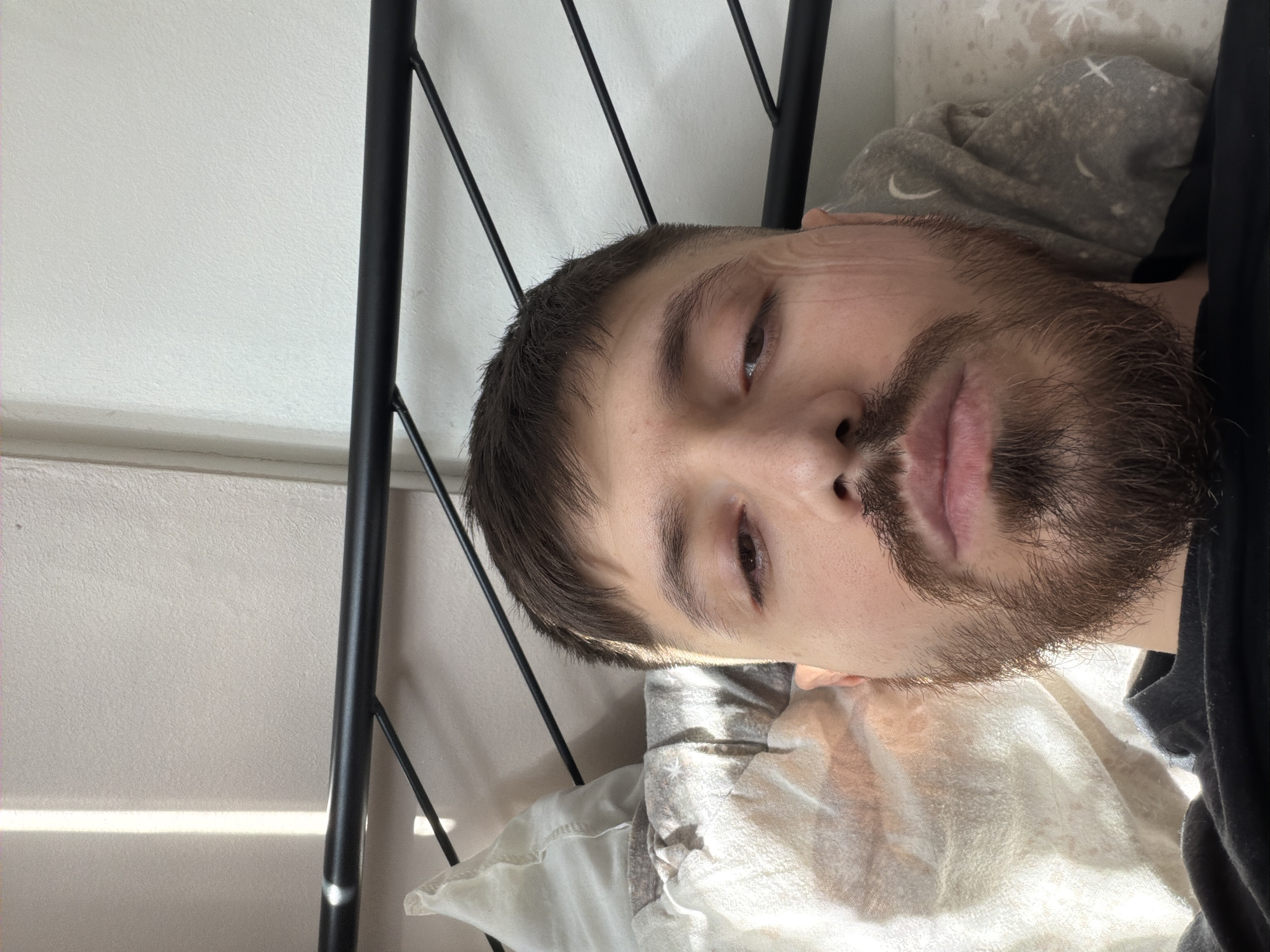 A man with dark hair and a beard lies on a white pillow in a bed with a black metal frame. The room appears to have white walls. The location cannot be determined as Pentre, United Kingdom without further information.