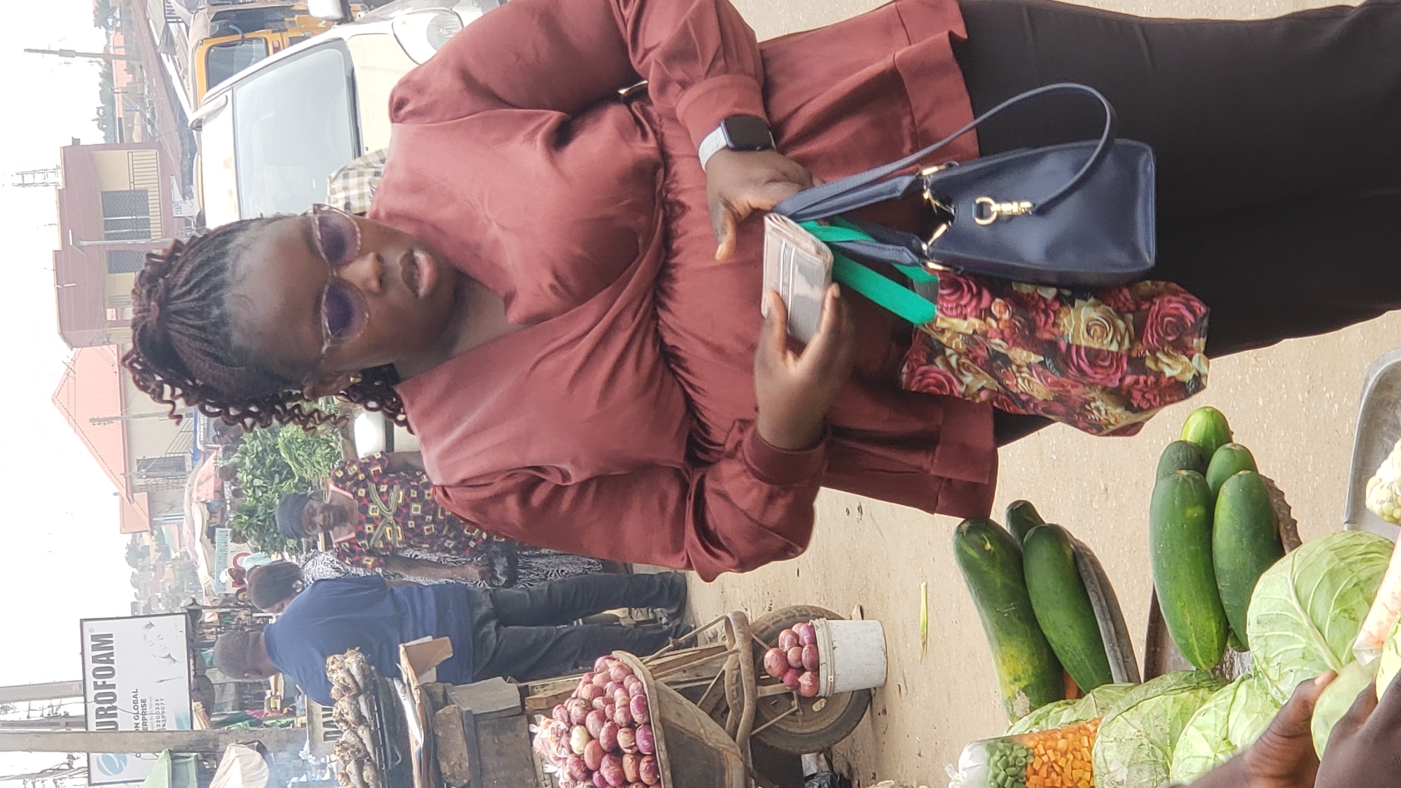 The image depicts a woman in Narayi, Nigeria, possibly at a market or roadside stall. She is wearing a maroon colored blouse and dark trousers. She has her hair in braids and is wearing sunglasses. She is holding a wallet or small purse in one hand and several bags hang from her shoulder including a navy bag and a floral printed bag.
In front of her are vegetables, including cucumbers and cabbage. Behind her is a wheelbarrow or cart loaded with red onions. There are other people in the background, along with a sign that says "UROFOAM." There are other stands with grilled fish and other objects that suggest a market setting. The environment is a busy outdoor street scene.