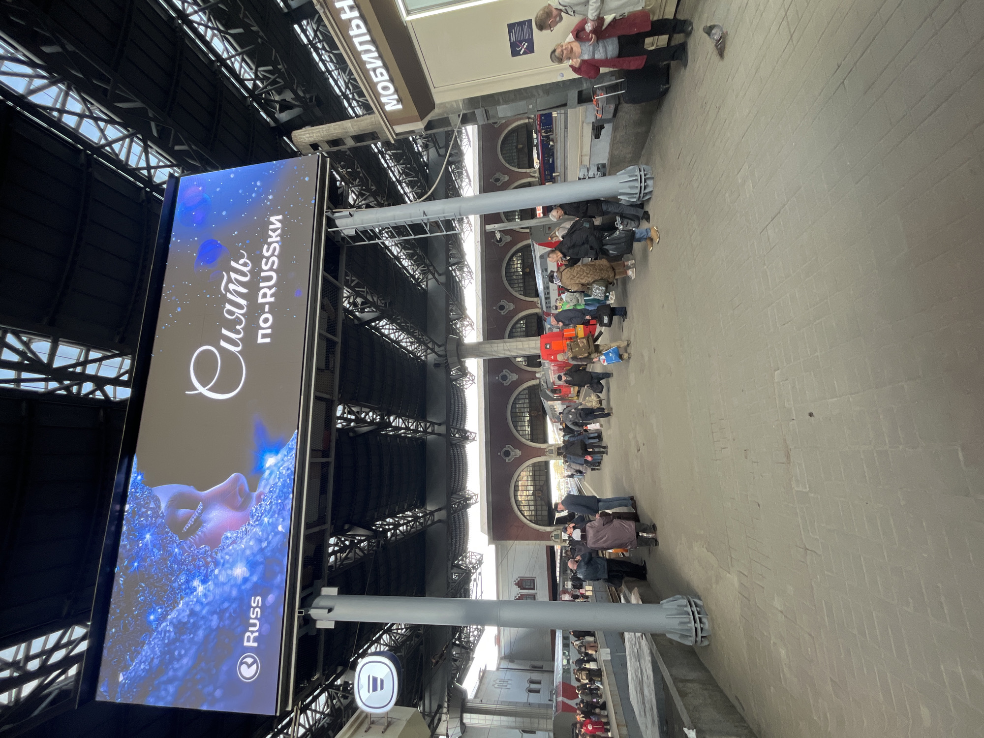 The image depicts a bustling scene at a railway station in Moscow, Russia.
**Location:** The image appears to be taken inside or near the entrance of a railway station in Moscow, Russia. This is indicated by the Cyrillic text on the advertisements and signs.
**People:** Numerous people are present, some appearing to be travelers with luggage. Several groups of individuals are standing around.
**Subjects/Objects:** A prominent digital advertising display is visible, featuring Cyrillic text that translates to "Dream in Russian." A sign above the display says "Мобильн" (Mobil'n) which translates to mobile. The railway station's architectural features include structural steel supports overhead and arched windows on the exterior walls.
**Actions/Interactions:** People are standing, moving, and possibly waiting for trains. There appears to be a queue of people lined up.
**Environment:** The scene is set in a large, indoor railway station hall with high ceilings and numerous architectural elements. The lighting appears to be a combination of natural daylight and artificial illumination. The station platform has been paved with light grey pavers.