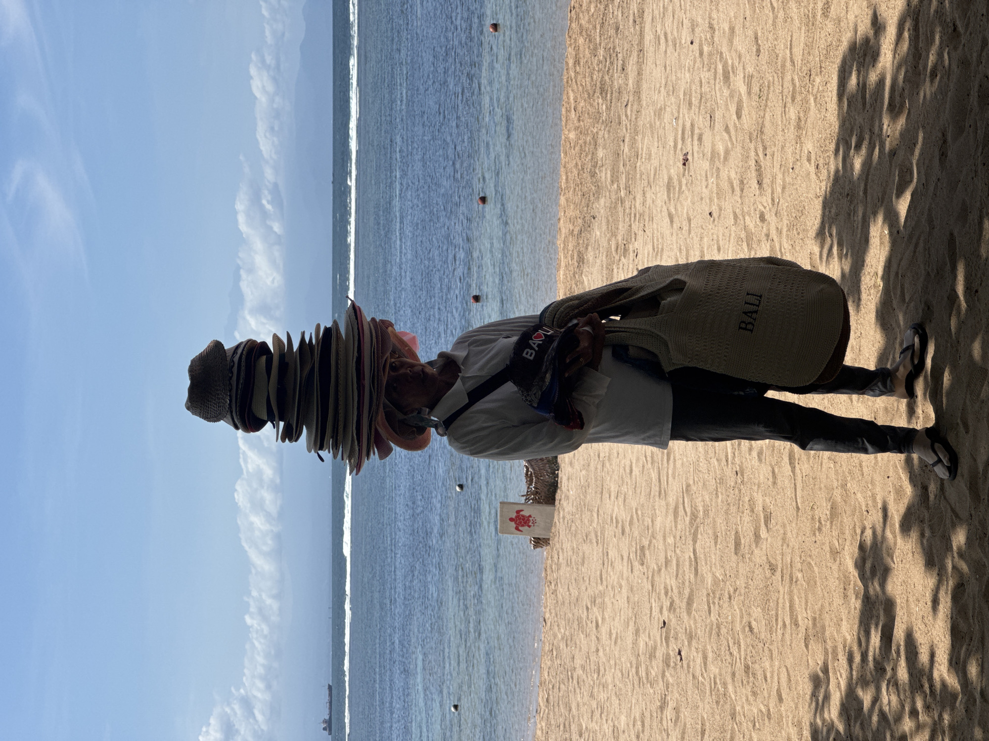The image shows a man standing on a beach in Kuta Selatan, Indonesia. He has a stack of hats balanced on his head. He is wearing a light-colored shirt and dark pants, and a bag with the word "BALI" on it is slung over his shoulder. A bag with the word "BAVU" is around his neck. The beach has light-colored sand and the ocean stretches to the horizon. A sign with a turtle is on the beach near the man. Some shadows are cast on the sand near the man's feet. The sky is blue with some clouds.