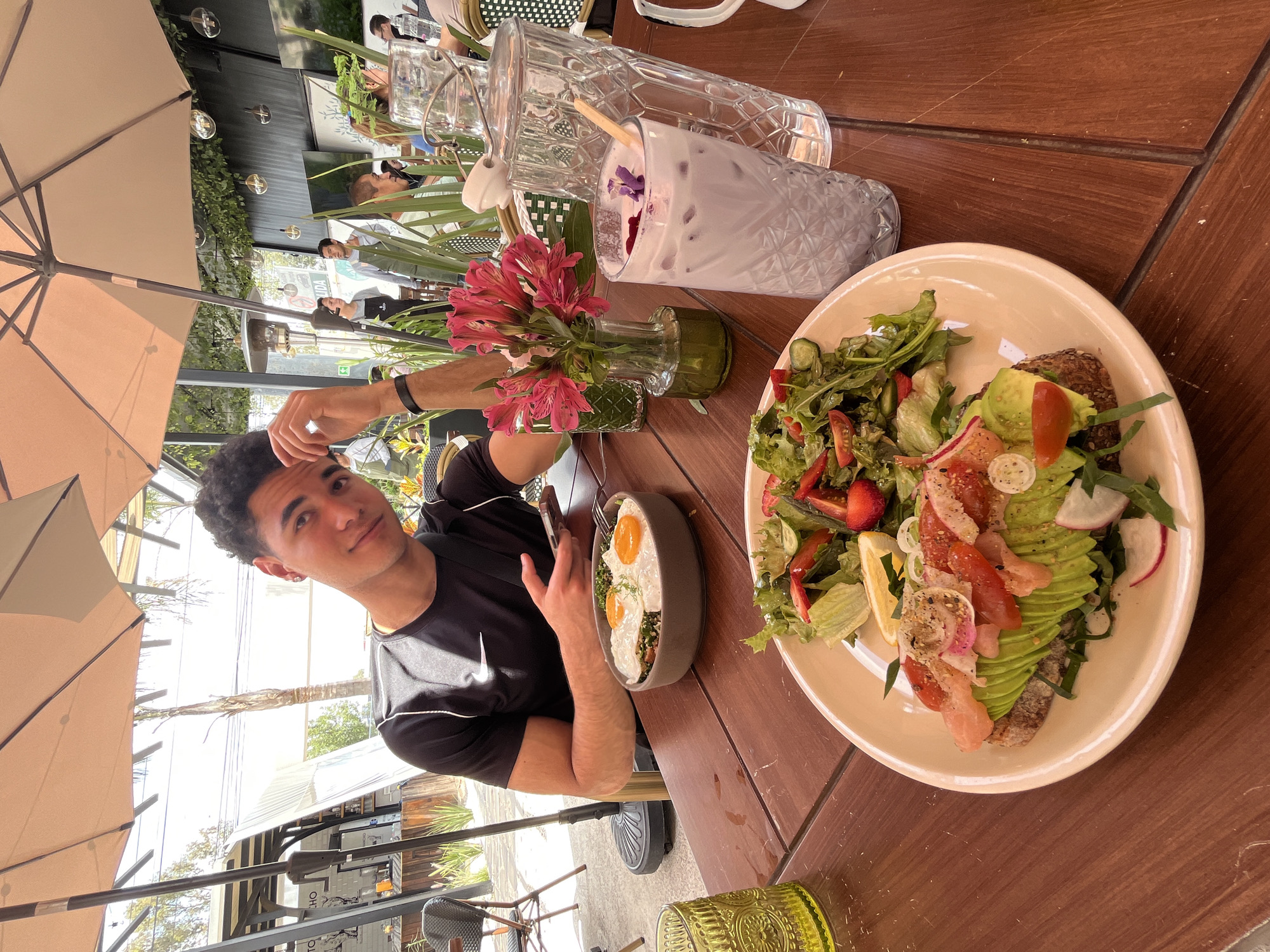 A young man sits at a wooden table at an outdoor restaurant in San Bernardino Tlaxcalancingo, Mexico. The table is covered with dishes: a salad with avocado and tomatoes on a beige plate, a bowl with two fried eggs, a glass of lavender-colored drink, a glass pitcher with water, and a small vase with pink alstroemeria flowers. The man is looking at the camera, his left hand on his head. An umbrella provides shade above him. Other people are seated at tables in the background, and greenery surrounds the restaurant.