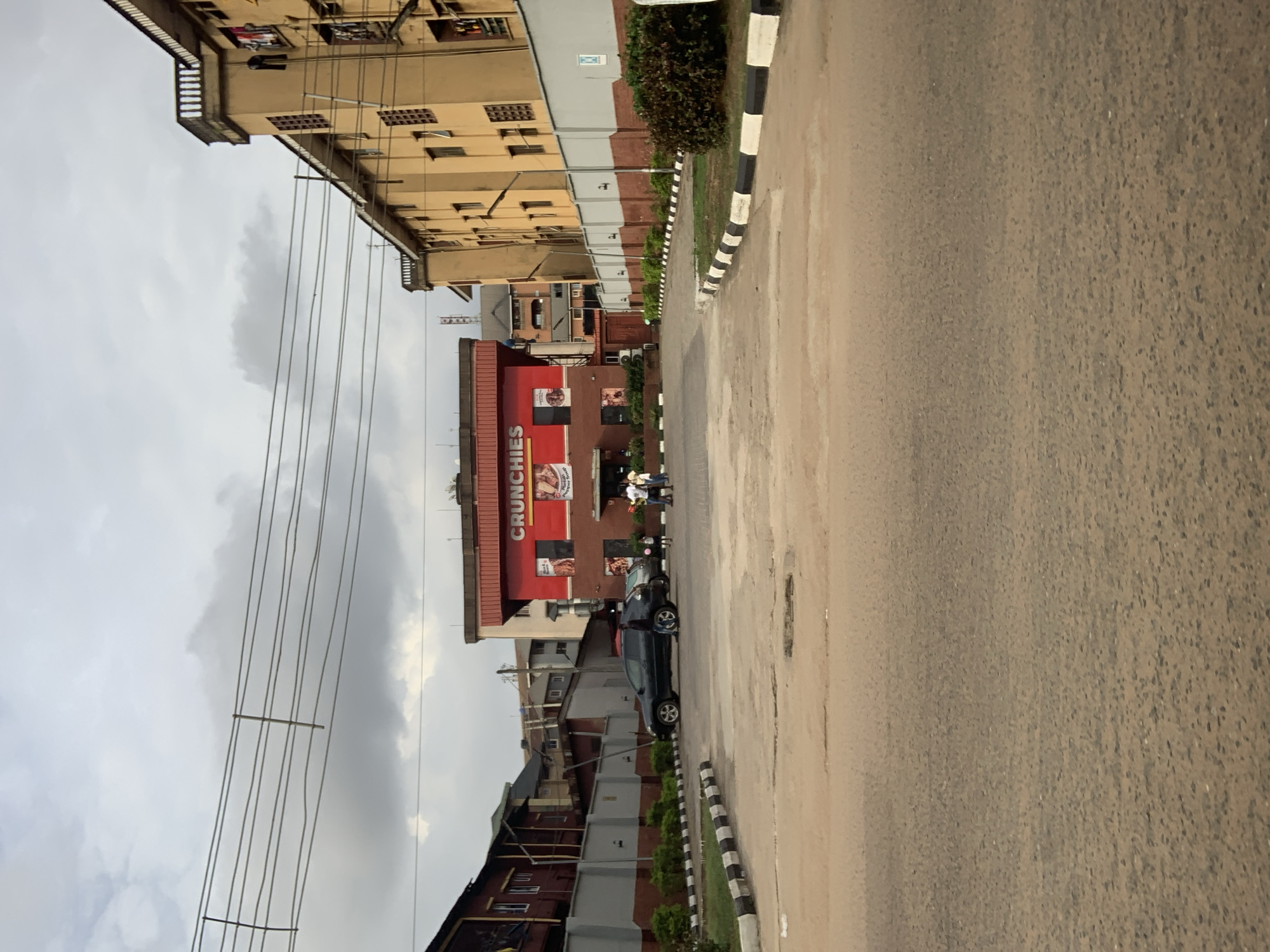 In Nkpor, Nigeria, a road leads towards a brightly lit red building with a sign that reads "CRUNCHIES." The building appears to be a fast-food restaurant. Several people are visible near the entrance. To the right of the restaurant, a tall, yellow apartment building stands with multiple windows and balconies. In front of the apartment building, a gray wall separates it from a grassy area. A dark-colored car is parked on the left side of the road, with a few people standing near it. Overhead, power lines crisscross the cloudy sky. The overall scene suggests a commercial and residential area on an overcast day.