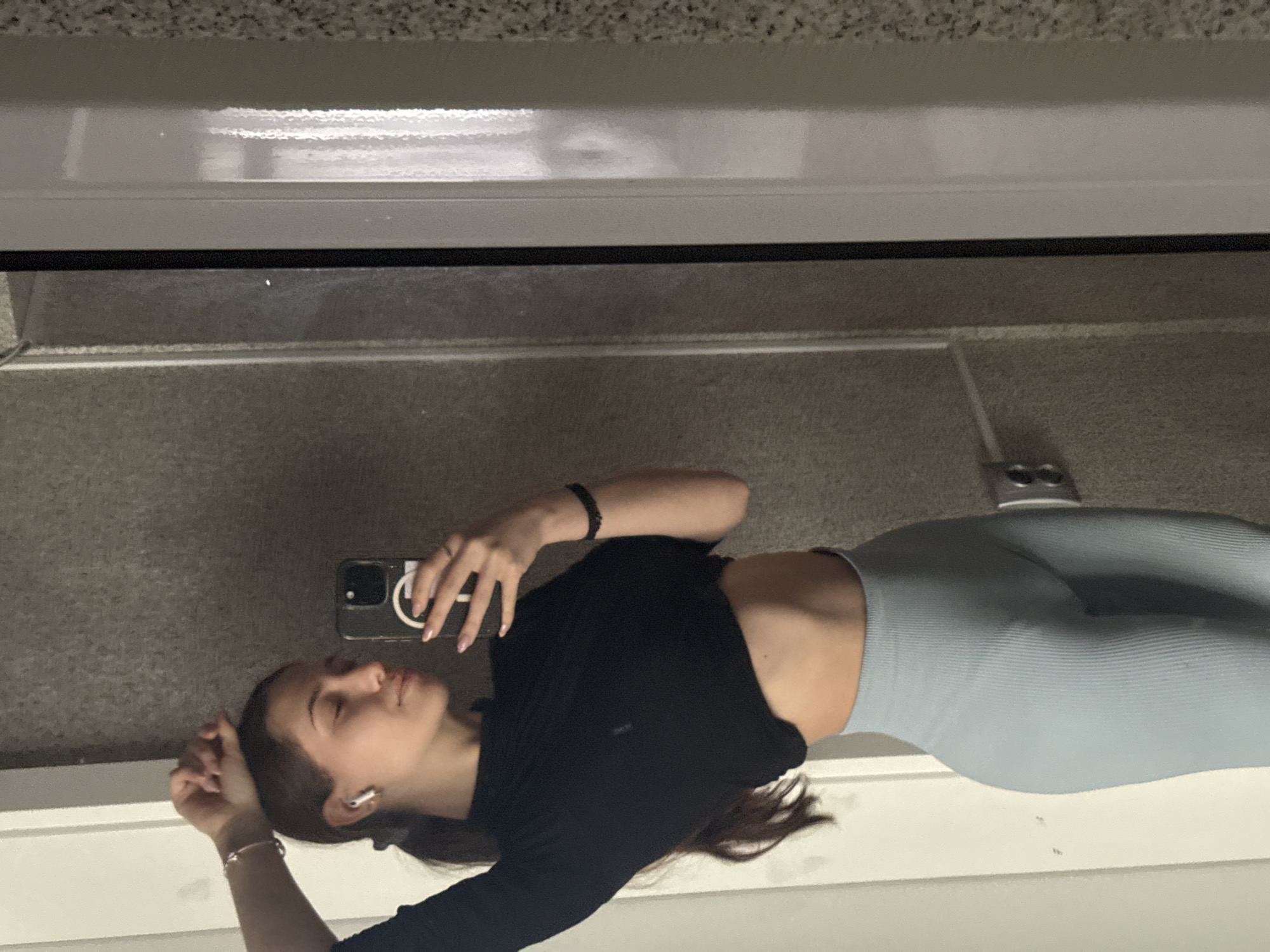 A young woman is shown from the waist up. She wears a black top and light blue leggings. She has dark hair and is wearing a bracelet, a black wristband, and an earpiece. She is holding a smartphone in her left hand. The background appears to be a mirrored wall. The image was taken indoors. The location is unknown, and there is no evidence to suggest it is in Sofia, Bulgaria.