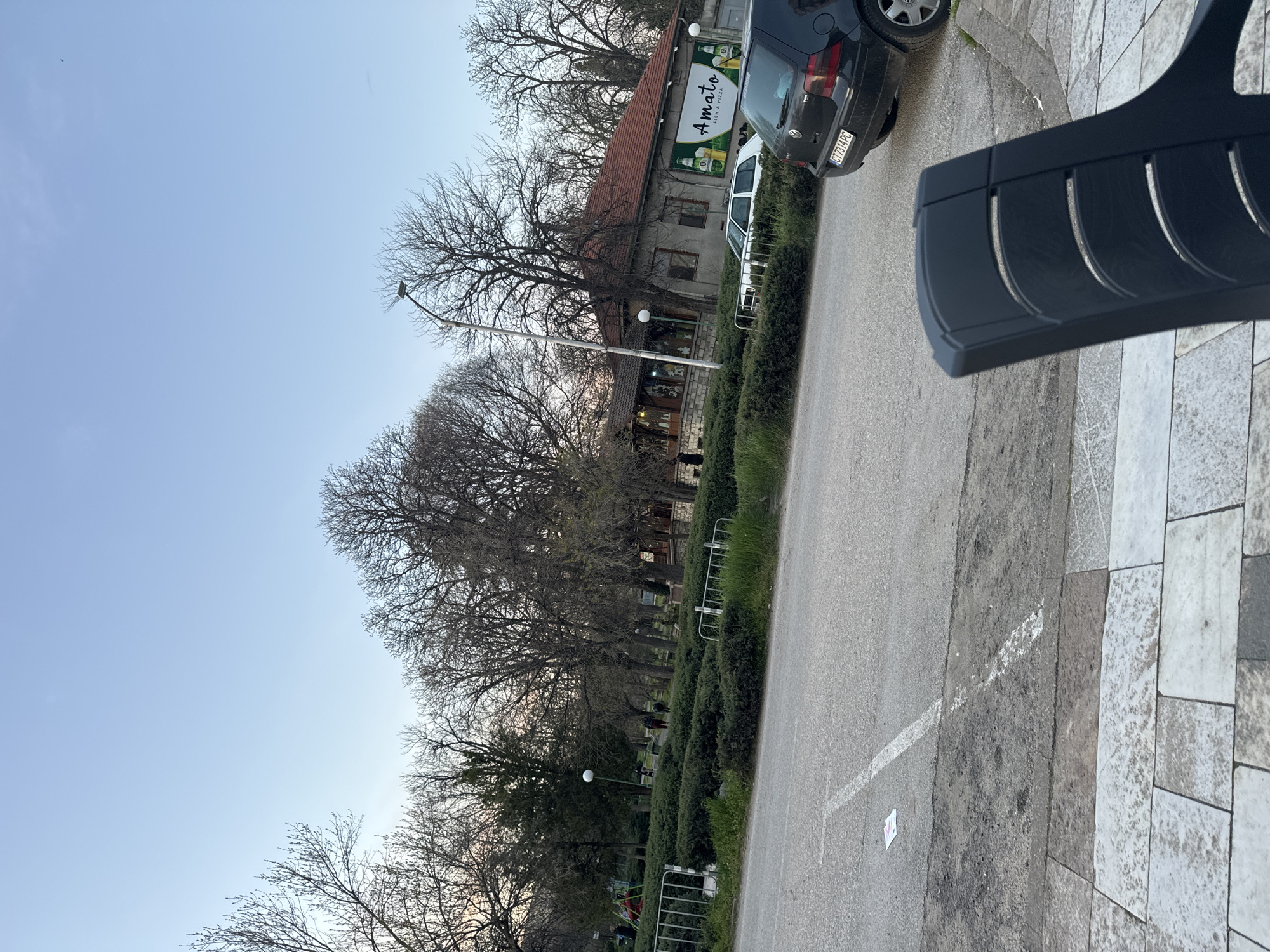 The image depicts a street scene in Chirpan, Bulgaria. A two-story building with a red roof is visible, featuring a sign for "Amato" (Pizza and Rum). Parked vehicles include a dark-colored Volkswagen Golf. A park/garden with bare trees and trimmed hedges runs along the side of the street. There's a sidewalk paved with stone tiles. Street lighting is also visible. The sky is light blue. In the foreground, the back of a dark bench is visible.