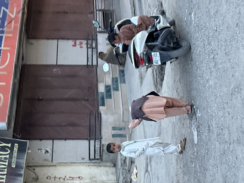 The image portrays an outdoor street scene in Quetta City Tehsil, Pakistan, during daylight. In the background, a building features two closed, dark brown roll-down metal shutters. Above the left shutter, a partial sign displays "PHARMACY FOR A BETTER LIFE," and a red sign with "SCHEZ" partially legible runs above the right shutter. Below the shutters, a set of concrete steps with alternating green and white risers leads to an elevated entrance, fronted by a black metal railing. An adult woman, dressed in a brown head covering and garment, is seated on these steps to the right.
In the midground, a white scooter is parked facing left, its rear wheel positioned within an indentation in the road. The scooter's rear license plate, "MAS-5022 QUETTA," is clearly visible. A young boy, wearing a brown shalwar kameez, is seated on the scooter, facing to the left of the frame.
In the foreground, the street surface is composed of rough, light grey asphalt with scattered small stones and debris. A young boy, dressed in a white shalwar kameez with green embroidered accents and brown sandals, stands on the left, looking directly at the camera with his hands in his pockets. To his right, another individual, clad in a peach-colored shalwar kameez and a dark brown vest, is bent over at the waist, facing away from the camera, with their attention directed towards the ground.