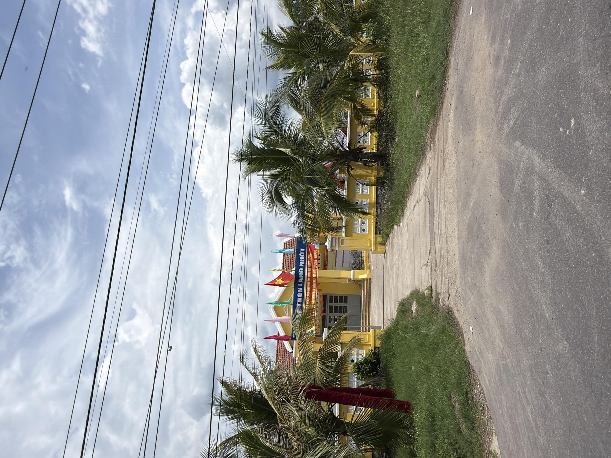 The image depicts the entrance to a yellow building in Diên Thọ, Vietnam, surrounded by palm trees and green grass. The building features a red-tiled roof and a blue sign with Vietnamese text. Several flags are displayed on poles above the entrance, including the Vietnamese flag. A pathway leads from a paved road to the building's entrance, which has double doors and a small set of steps. The scene is captured on a cloudy day, with power lines visible against the sky. The area appears to be a public space, possibly a community center or government building.