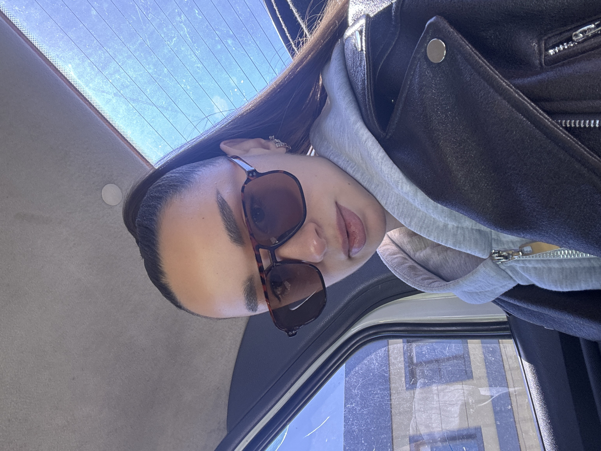 A woman with long brown hair is wearing brown sunglasses, a grey hoodie, and a black leather jacket. She is sitting inside a vehicle with a window showing a building. It is impossible to determine the specific location in Belorechensk, Russia, without further information.