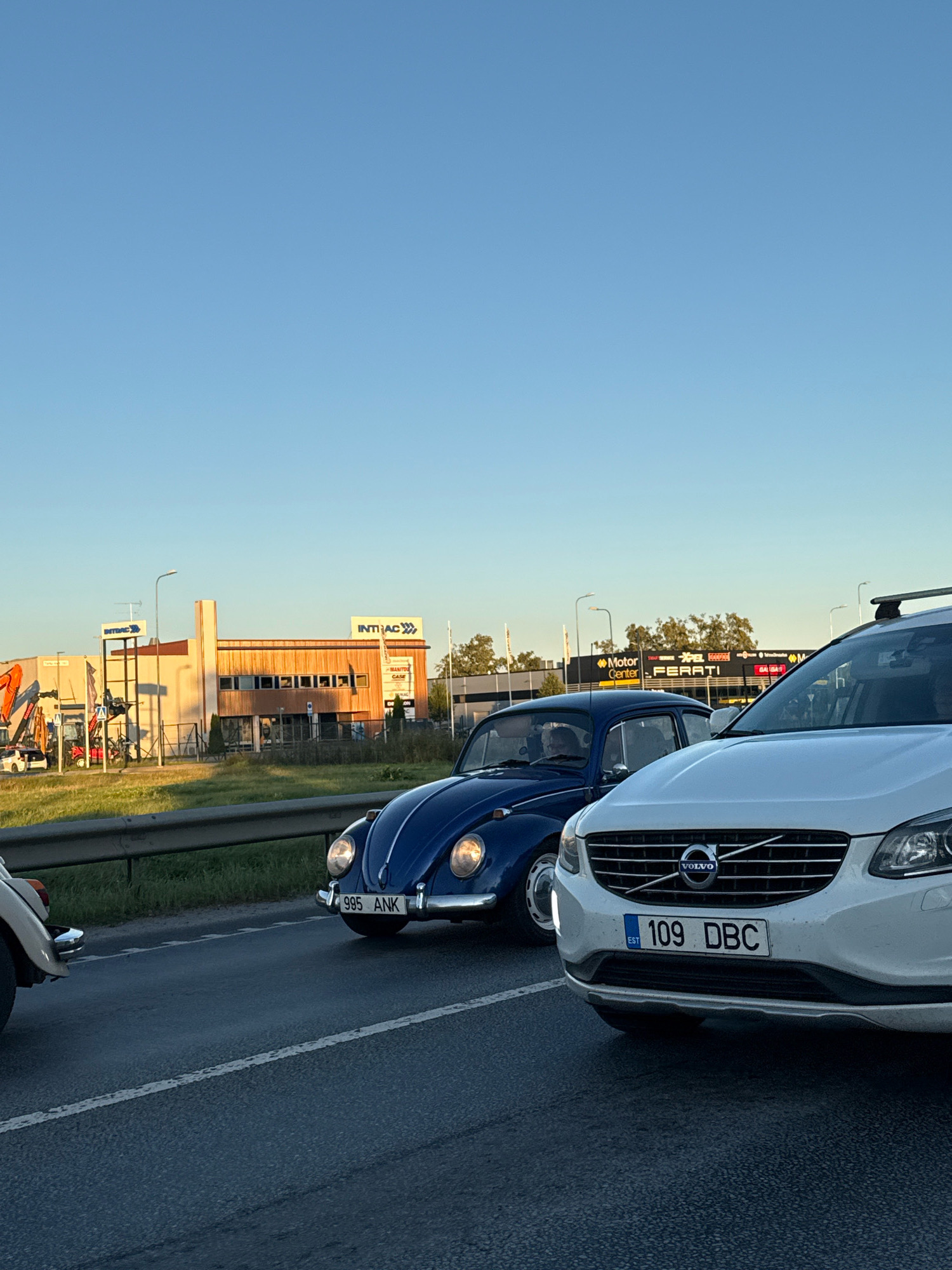 The image shows a street scene in Peetri alevik, Estonia, with several cars. In the foreground, a white Volvo SUV is parked on the right side of the frame, and a blue Volkswagen Beetle is in the left lane. Behind the cars, there is a building with "INTRAC" signage and a lot with construction equipment. The sky is clear and blue, suggesting it is daytime. The overall impression is that of a typical day in a suburban area.