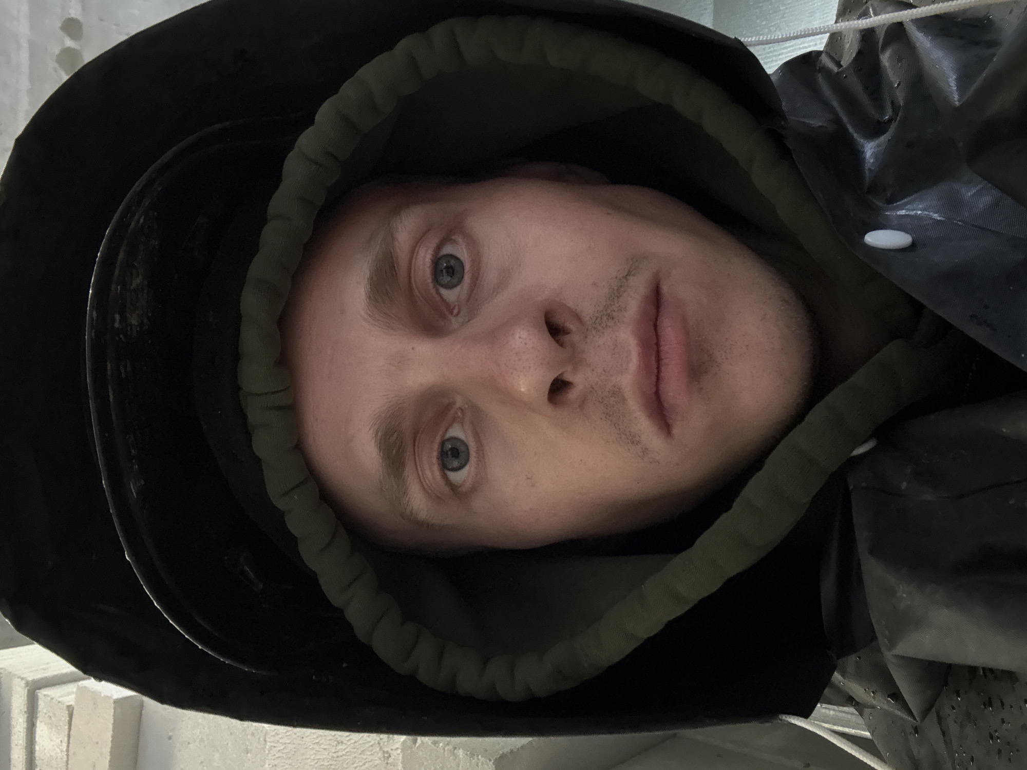 The image is a close-up, front-facing selfie of a person wearing a black hood and a dark-colored, possibly waterproof, jacket. The person has light skin, light-colored eyes, and short, light-brown hair that is mostly obscured by the hood. They have a light mustache and stubble. The jacket appears to be wet, with visible water droplets. A portion of what looks like a brick wall is visible in the upper left corner. The person is indoors, suggested by the lighting and the background texture. No specific Moscow landmarks or identifiable city elements are present in the image.