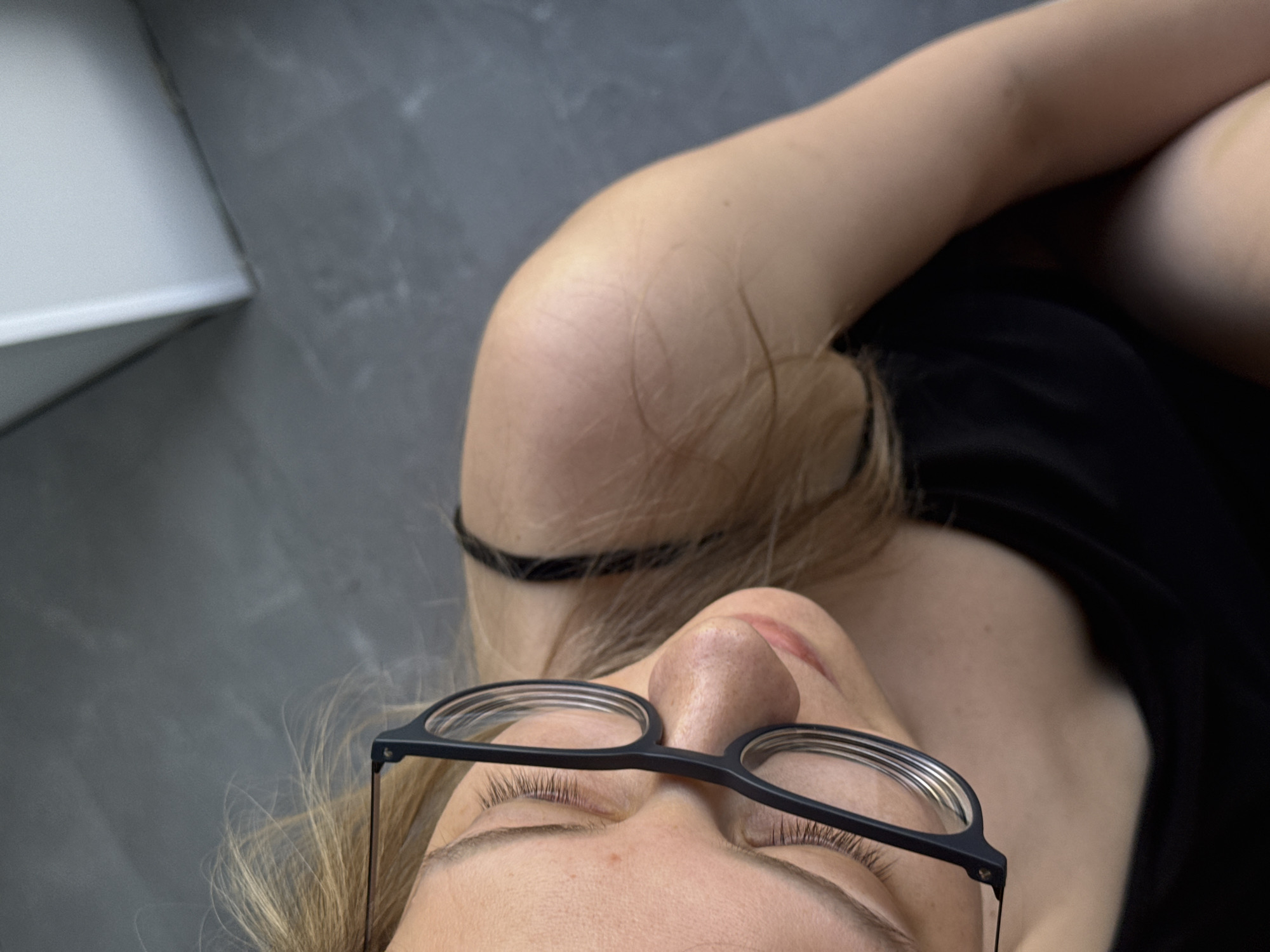 A top-down close-up photograph shows a fair-skinned person with closed eyes, wearing eyeglasses with thin black frames. The person's nose is prominent. The person is wearing a black top. The background consists of a gray floor and part of a white object, possibly furniture. The location and date are not provided. There is no information to suggest this image was taken in the city of Chaikovsky, Russia.