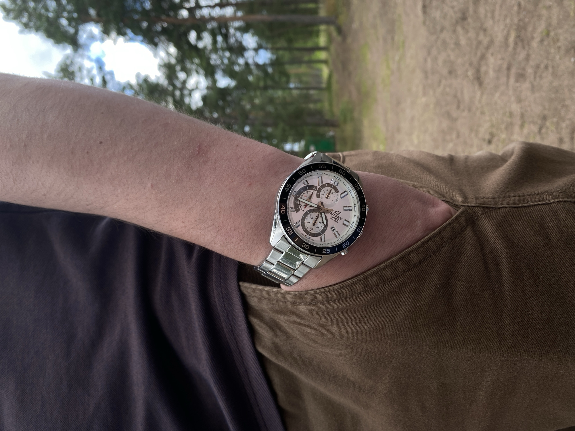 A person's arm wearing a silver watch with a white face and multiple subdials is positioned with their hand in the pocket of brown pants. The person is wearing a dark t-shirt. The background is a blurred forest with tall trees and a bright sky. The scene appears to be outdoors in a natural setting, possibly a park or wooded area. The location is Baltiiskoe, Russia.