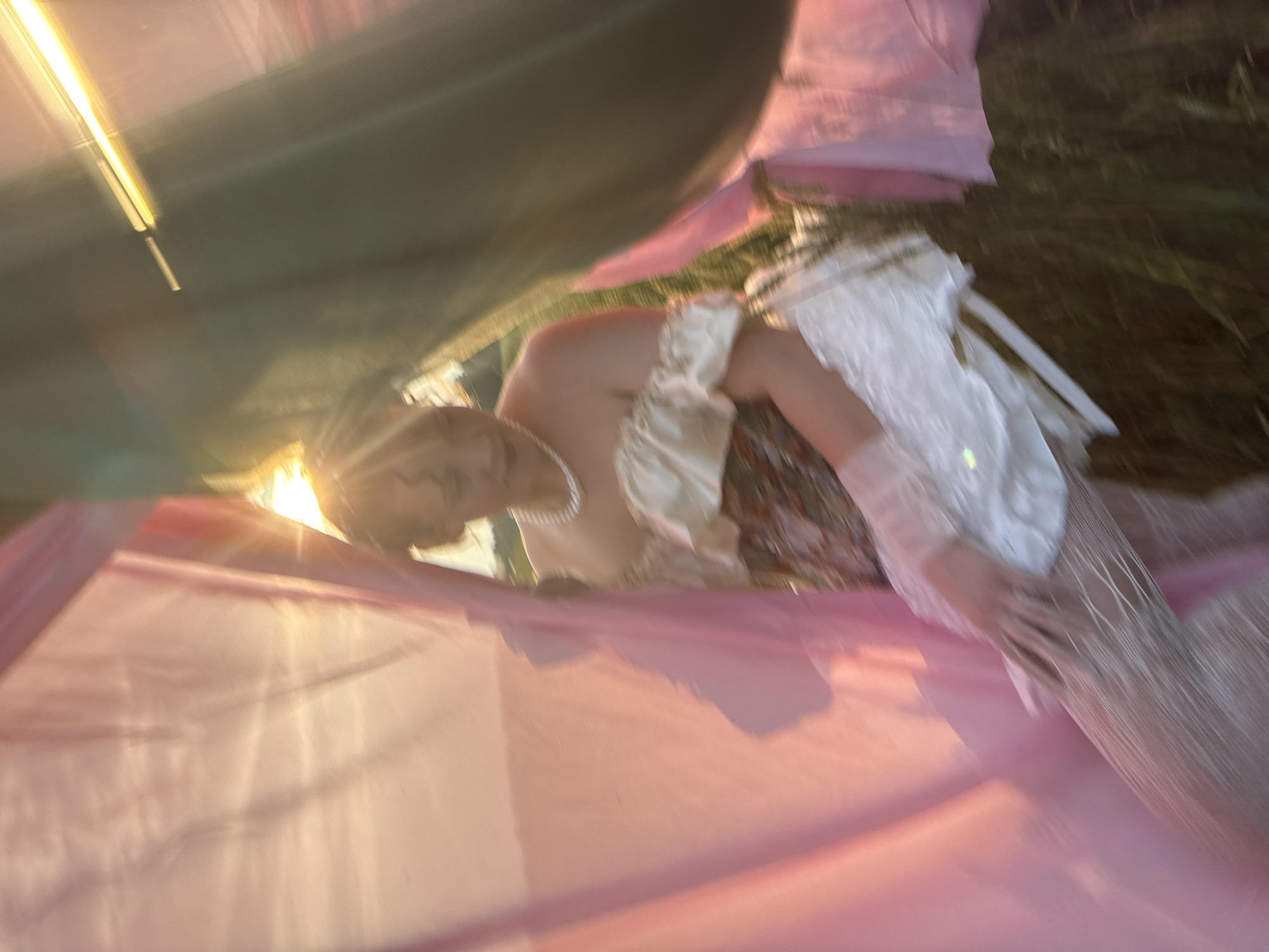 In Malyi Bukor, Russia, a person is seated amidst flowing fabrics of pink and green. The setting appears to be outdoors, with tall grass visible in the background and the sun setting, casting a warm glow. The individual is wearing a dress with an off-the-shoulder design, adorned with a pearl necklace. The overall image has a dreamy, somewhat blurry quality, suggesting movement or a soft focus.