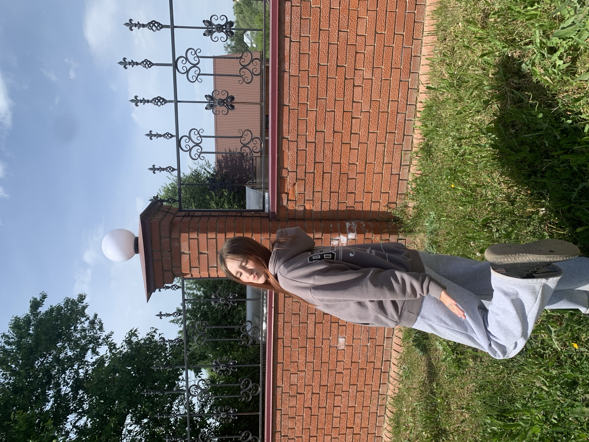 A medium-level outdoor shot depicts a young woman standing on a grassy area adjacent to a brick wall and a black wrought iron fence.
The woman is dressed in a grey hooded sweatshirt and light grey sweatpants. Her pose appears relaxed.
The brick wall is red and appears to be part of a more extensive structure. A section of a black wrought iron fence runs parallel to the wall.
The background includes trees and a partially visible building. The sky is blue with scattered clouds.
The location is specified as Фоки, Russia.