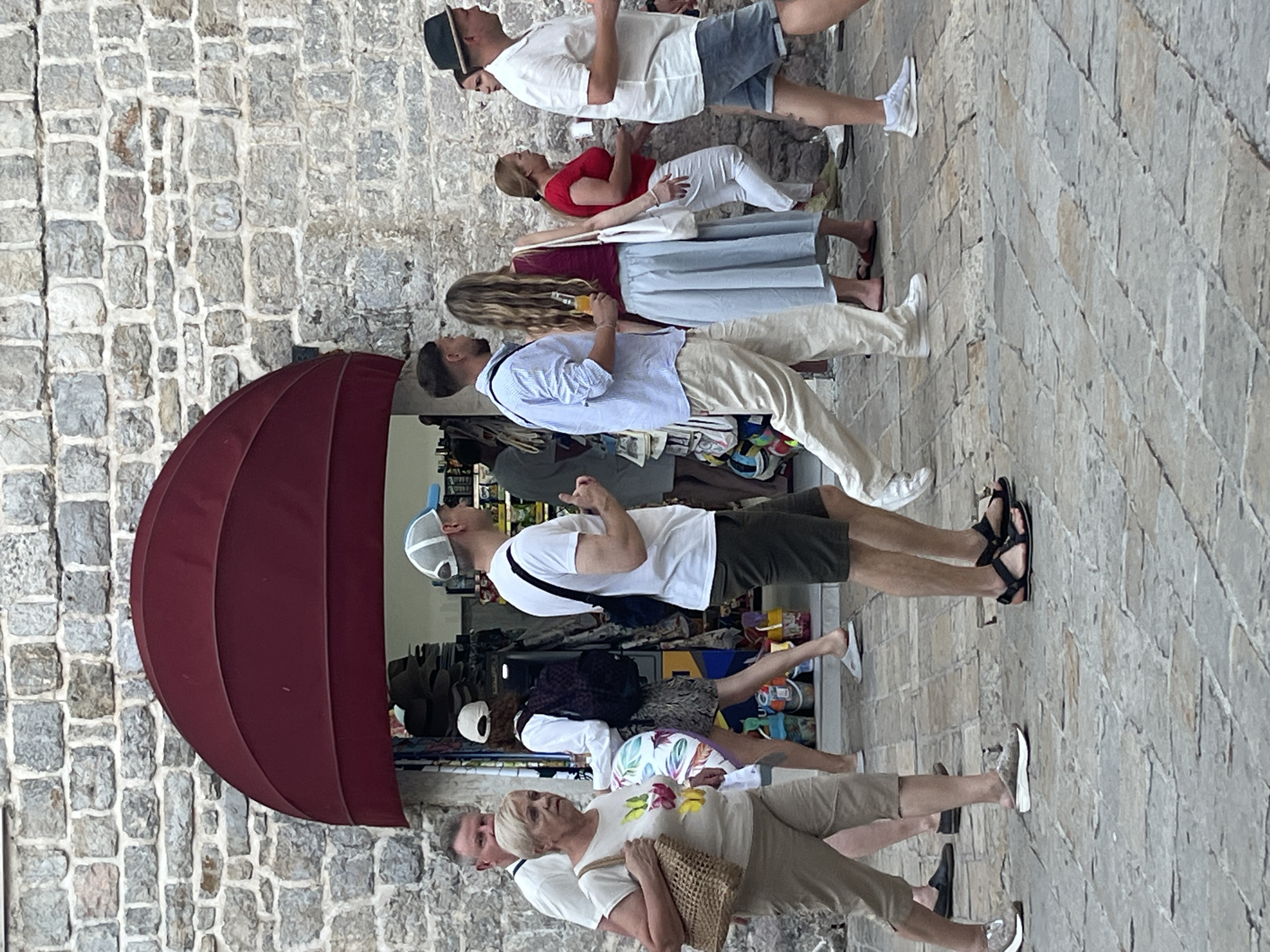 The image shows a group of people walking on a cobblestone street in Budva, Montenegro, in front of a stone building with a red awning. The building appears to be a shop selling souvenirs or clothing. The people are casually dressed, and most are walking towards the right of the frame. One woman in the foreground is carrying a woven bag and wearing a t-shirt and pants. To her right, a man in a white t-shirt and khaki shorts is walking. Further back, a man in a light blue striped shirt and beige pants is walking with a woman in a red top and a light blue skirt. Another woman in a red top and white pants is walking. On the far right, a man in a white shirt and blue shorts is walking. The overall scene depicts a typical day in a tourist area.