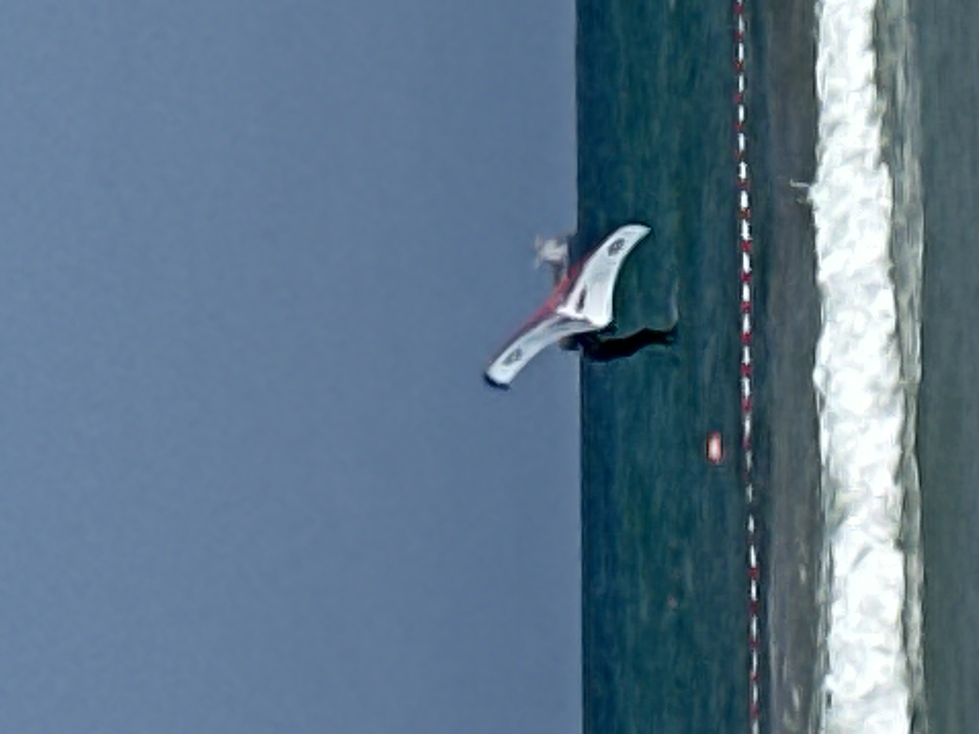 A person is wing foiling in the Mediterranean Sea off the coast of Kiryat Yam, Israel. The person is wearing a dark wetsuit and holding onto a white and red wing. They are positioned in the water, with the wing extended above them, appearing to be in mid-air or about to take off. The sea is a deep blue-green color with white waves crashing on the shore in the foreground. A red and white rope barrier is visible along the shoreline. The sky is a muted blue, suggesting it is overcast or dusk. In the background, a small vessel with a white structure on its deck is visible on the horizon.