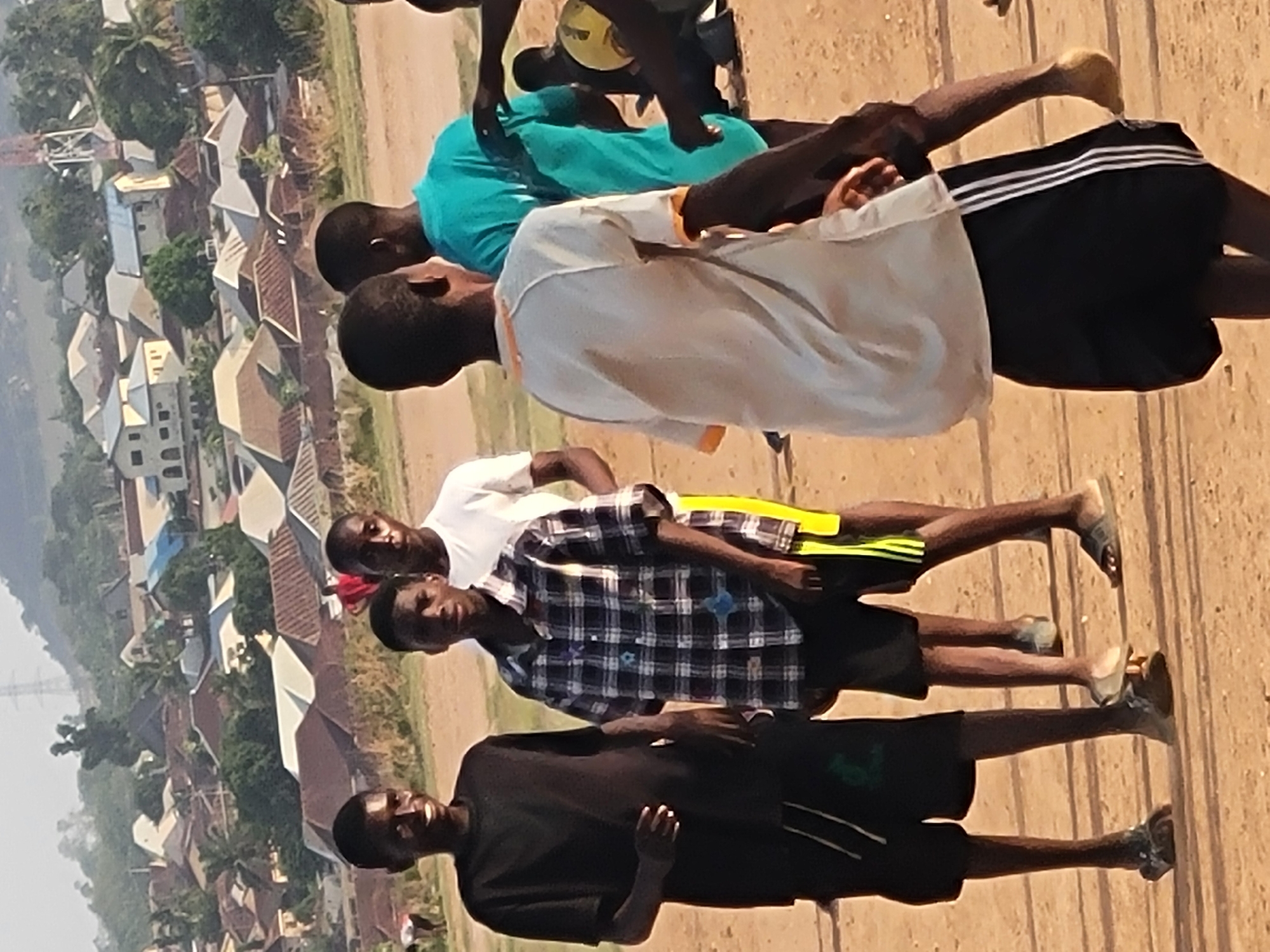 A group of four young men stand outdoors in Ibo, Nigeria. They are facing forward, arranged in a line, with the background displaying buildings and a rural cityscape.
The first individual is wearing a black shirt and dark shorts with a white stripe, and sandals on his feet. The next person is clad in a blue and white plaid shirt with yellow striped shorts and dark sandals. The third figure has a white shirt and black shorts. The fourth individual wears a light blue shirt with black shorts bearing white stripes. The first three are barefoot. All four subjects appear to be young men. The setting suggests a rural or semi-rural region within the city.