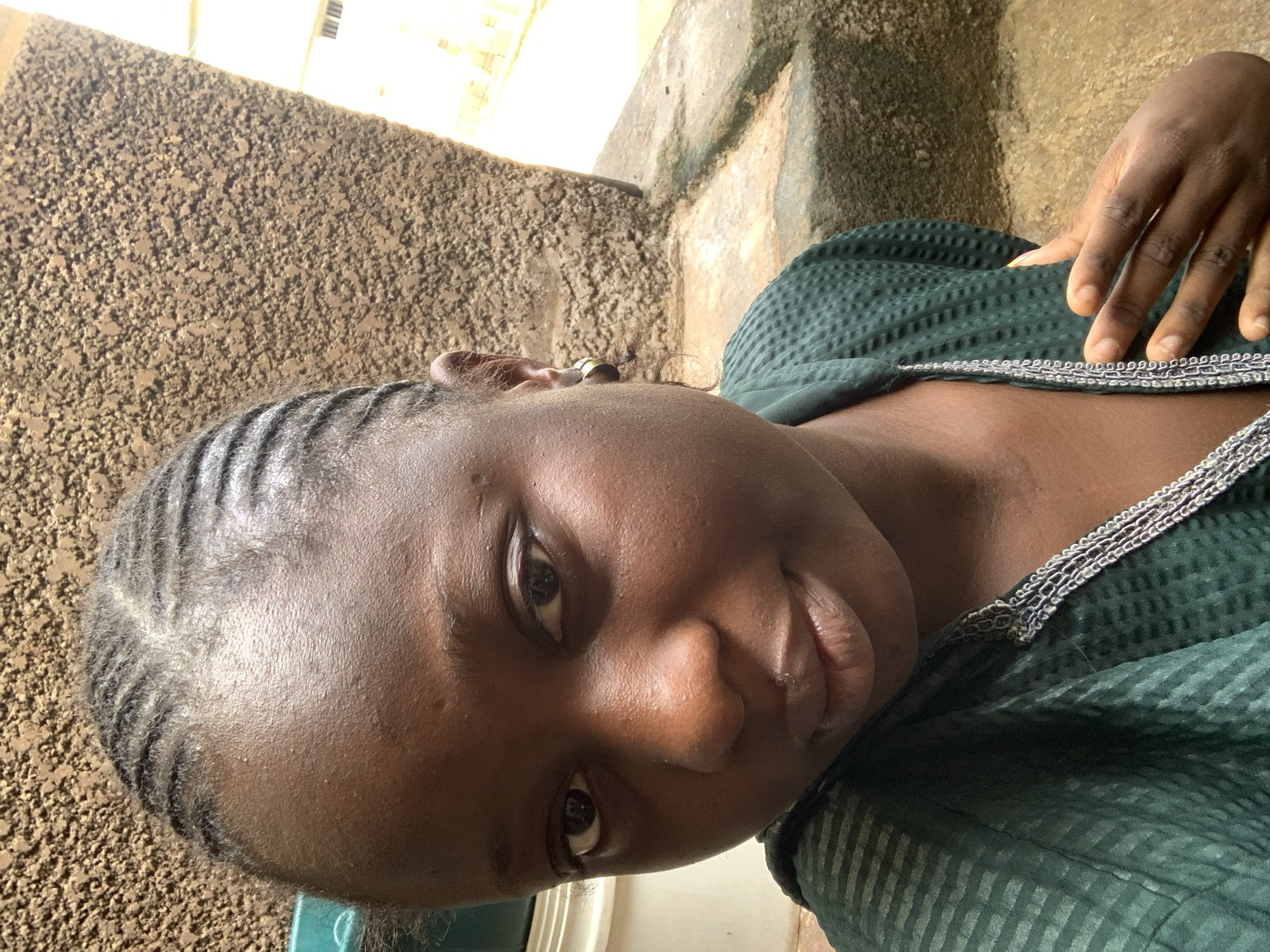 A medium shot shows a young Black woman from the chest up, looking directly at the camera. She has dark skin, braided hair, and is wearing a dark green patterned top with a silver trim around the neckline. A gold hoop earring is visible in her left ear. Her right hand is resting on her chest. The background is out of focus and appears to be an outdoor setting with a textured, brown wall and some concrete steps. The lighting suggests it is daytime.