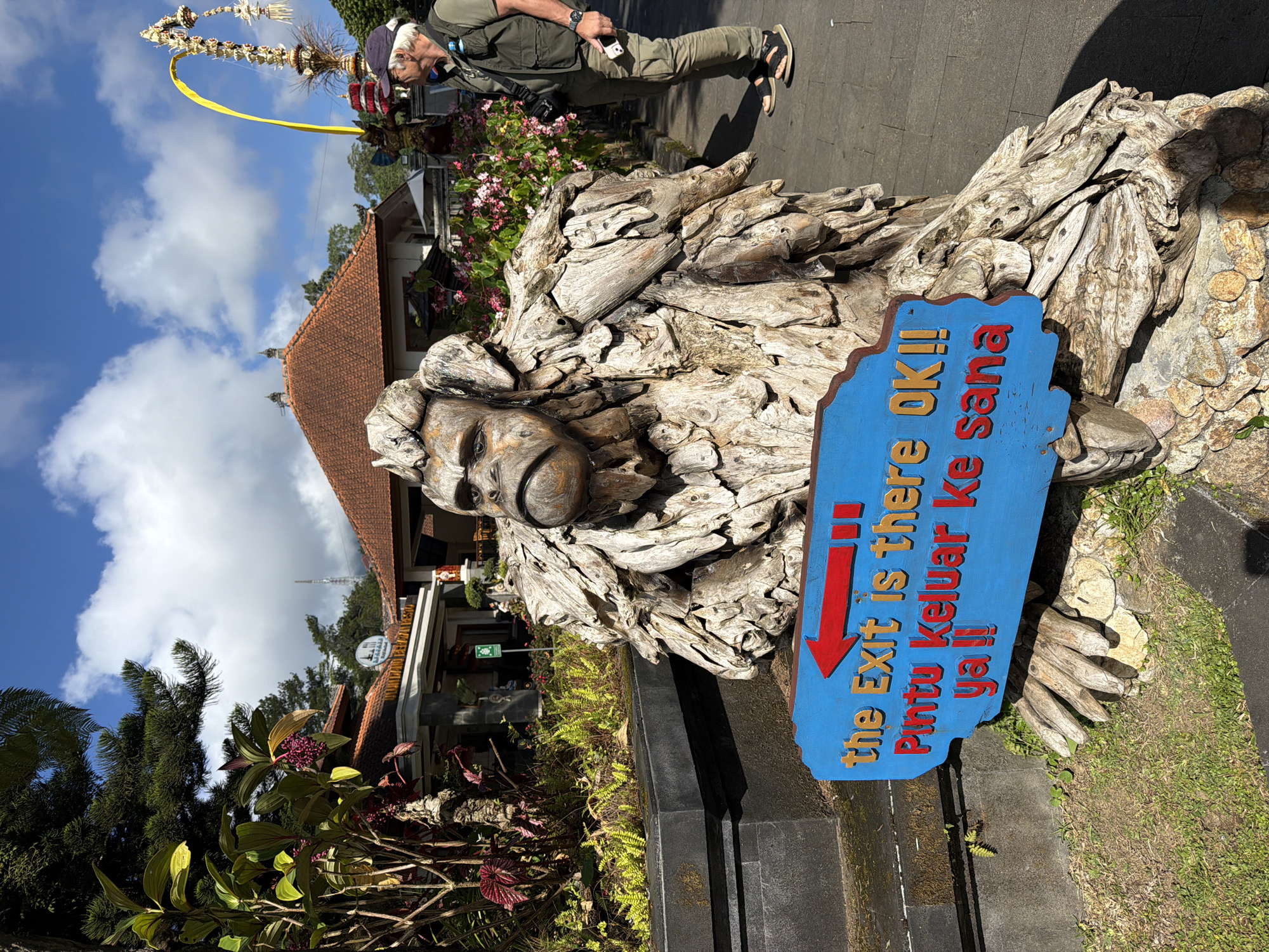 A wooden statue of a monkey-like creature stands in front of a building in Baturiti, Indonesia. The statue is made of driftwood and has a sign attached to it that reads "the Exit is there OK!! Pintu keluar ke sana ya!!". A man wearing a green vest and a blue cap is standing behind the statue, looking to the right. The building in the background appears to be a restaurant or a shop. The sky is blue with some clouds. The ground is paved with gray stones.