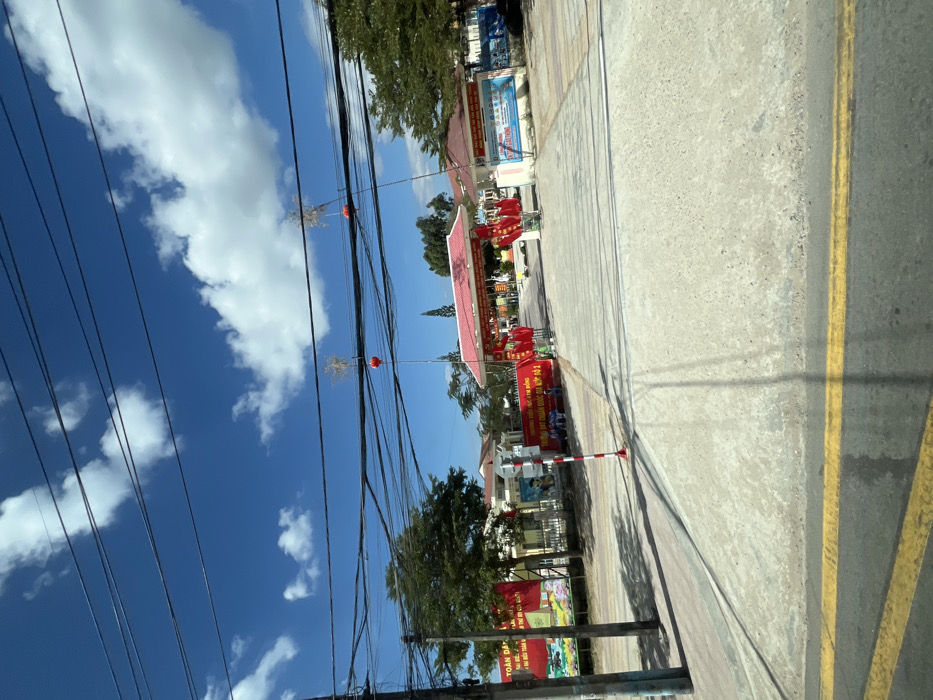 A street scene unfolds beneath a bright blue sky dotted with white clouds. Utility lines crisscross the sky, adorned with occasional red spheres. Banners and flags decorate buildings lining the street. A red-roofed structure dominates the scene, possibly a town hall or community center, marked with festive decorations. The paved road is marked with double yellow lines.