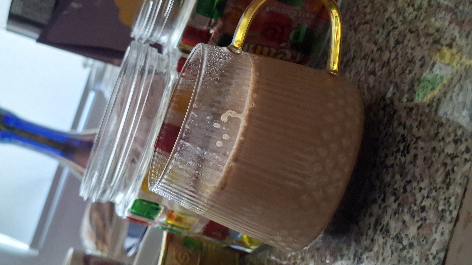 A glass mug filled with a light brown beverage sits on a speckled granite countertop. The mug has vertical ridges and a golden handle. The beverage has a frothy top with small bubbles. In the background, out of focus, are two glass jars with colorful contents, possibly candy, and a blue vase or bottle. The scene is indoors, with a hint of natural light coming from a window in the background. There are no people visible in the image. The location is stated to be Myadzyel, Belarus.